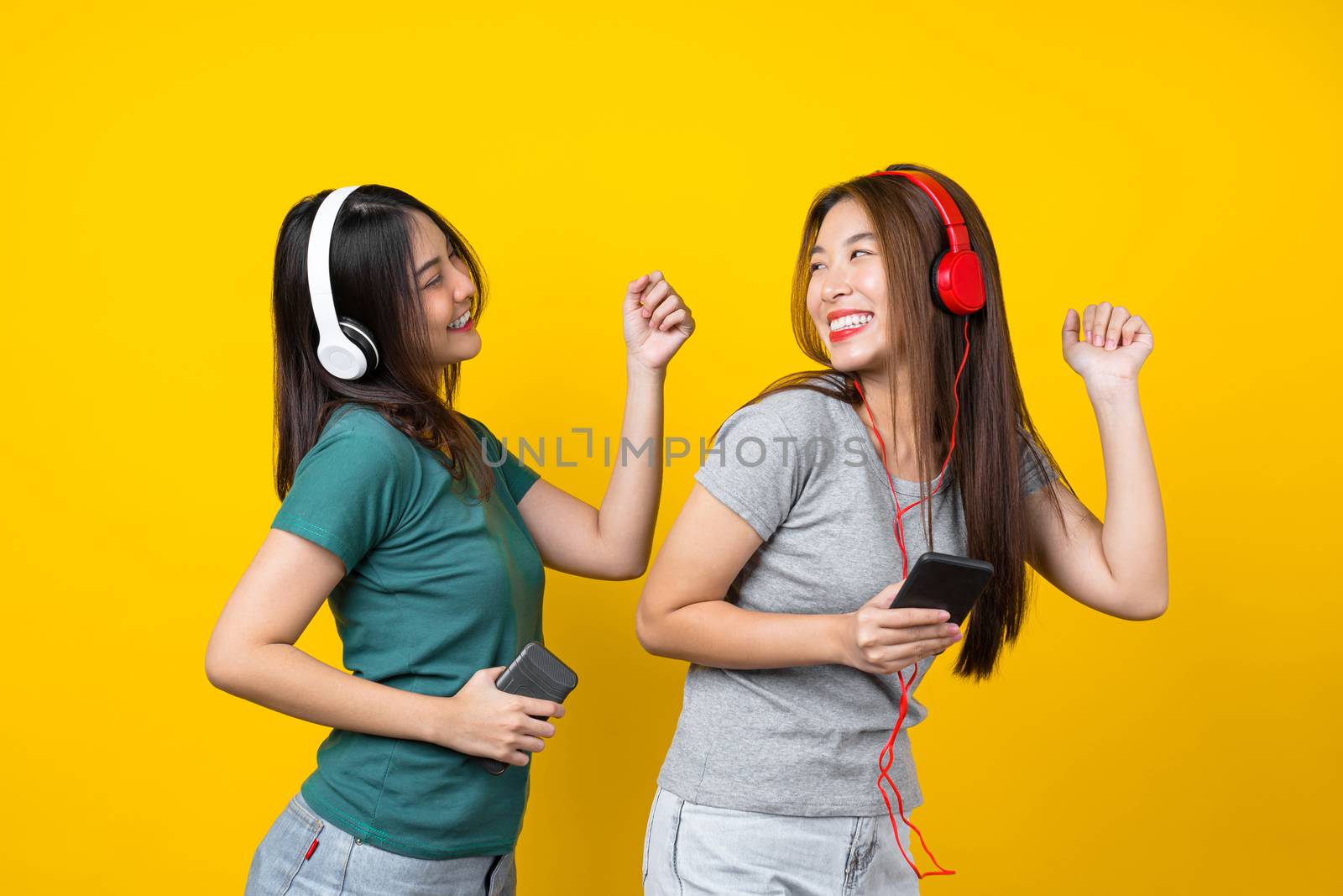 Two Happiness Asian smiling young woman wearing wireless headphones for listening music via smart mobile phone and dancing on isolated yellow color background, Lifestyle and leisure with hobby concept