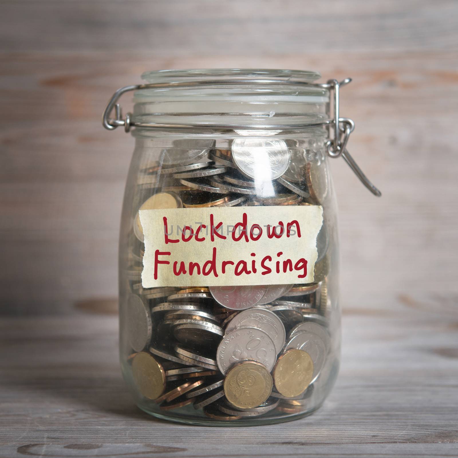 Coins in glass money jar with lockdown fundraising label. Vintage wooden background with dramatic light.