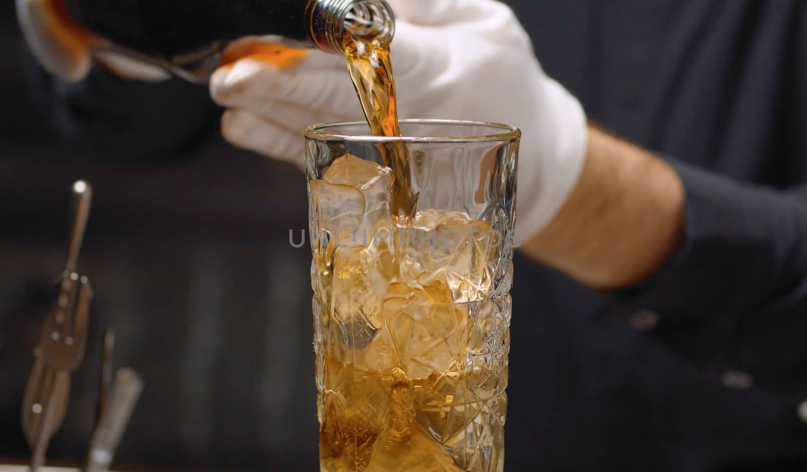 Making a Long Island Ice Tea Cocktail. Close up bartenders hands pouring cold tea into a crystal glass with ice.