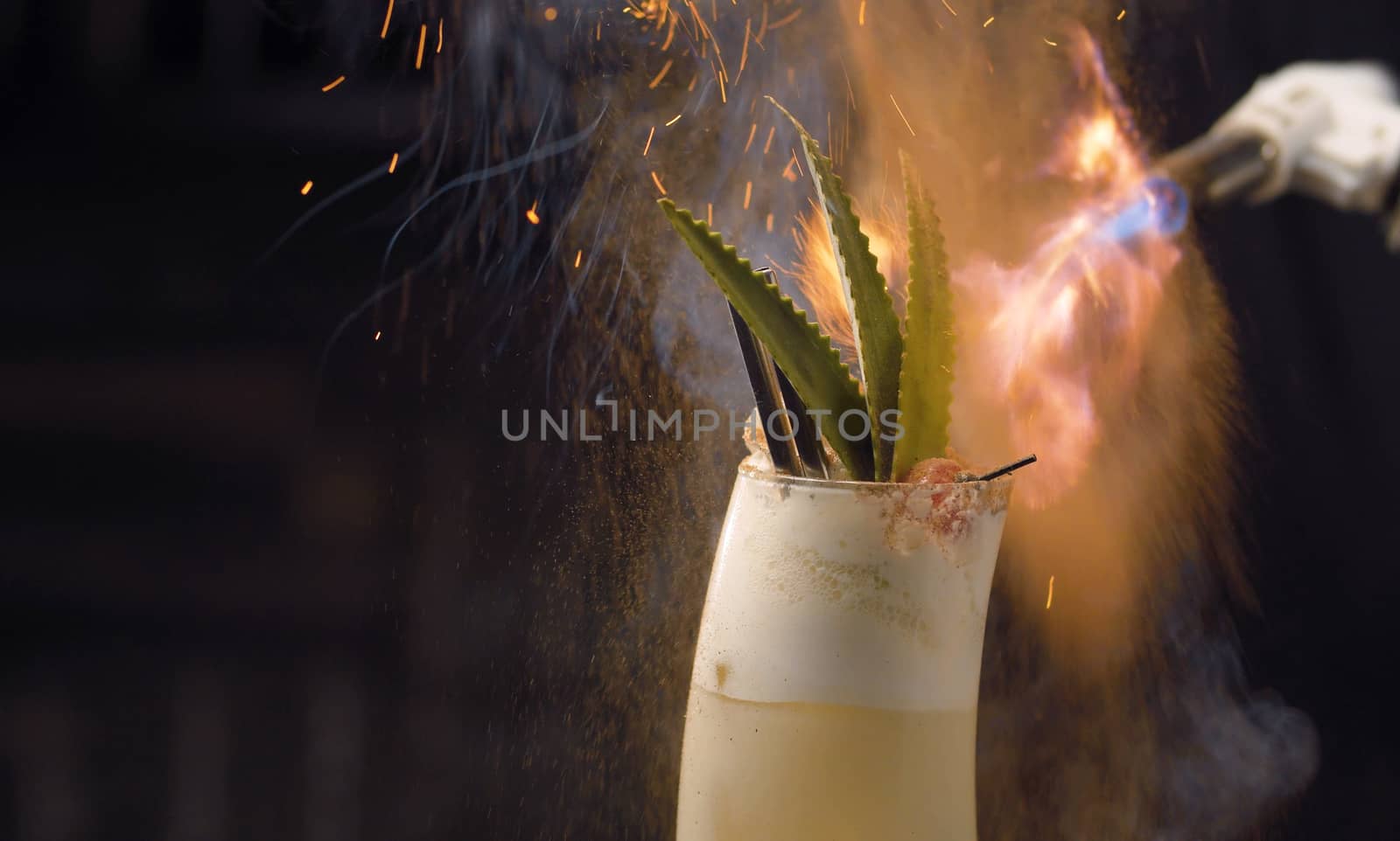 Close up bartenders hands sprinkling with burning cinnamon pina colada cocktail. Glass on the background of the fire. Bar drinks series.