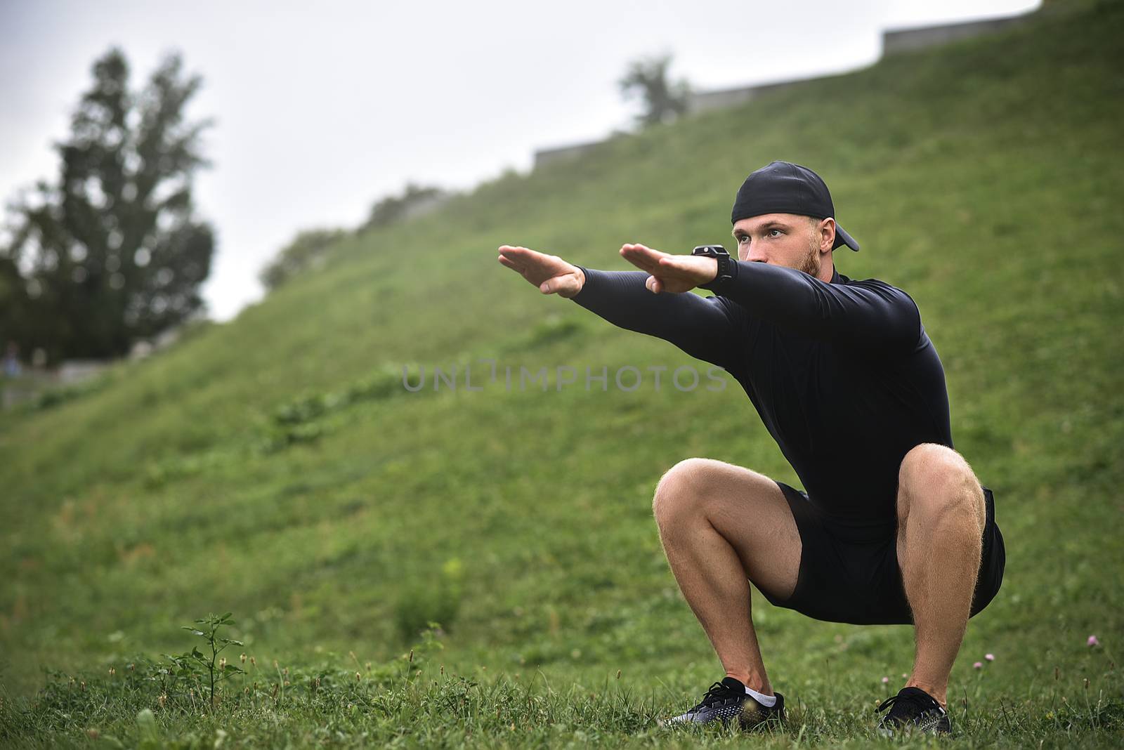 Young caucasian man performing squats before jogging on footpath in park.