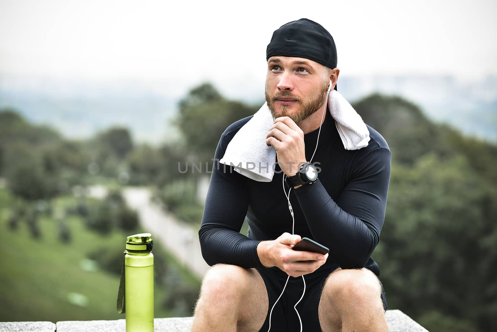 Muscular bearded athlete with towel checking burned calories on smartphone after good workout session on city park.