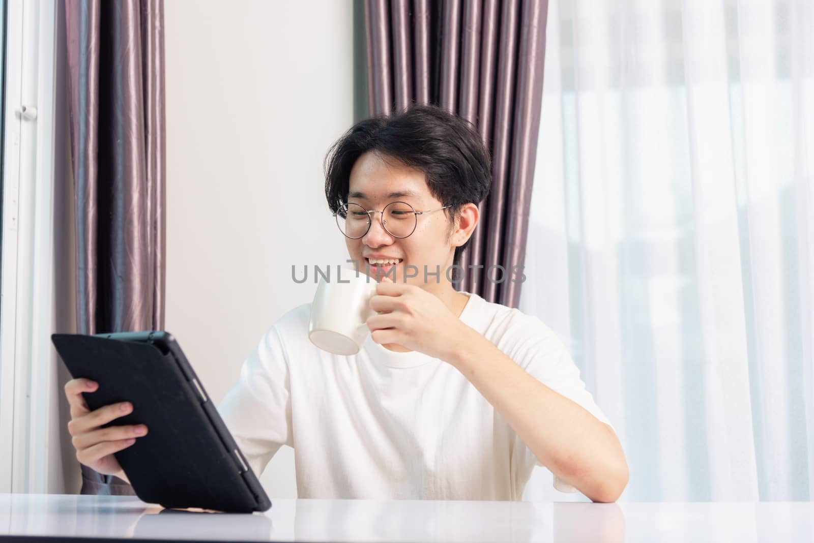 Happy Asian young business handsome man work from home office wear glasses, t-shirt comfortable he smiling and using a black modern smart digital tablet computer to read email or learning on desk