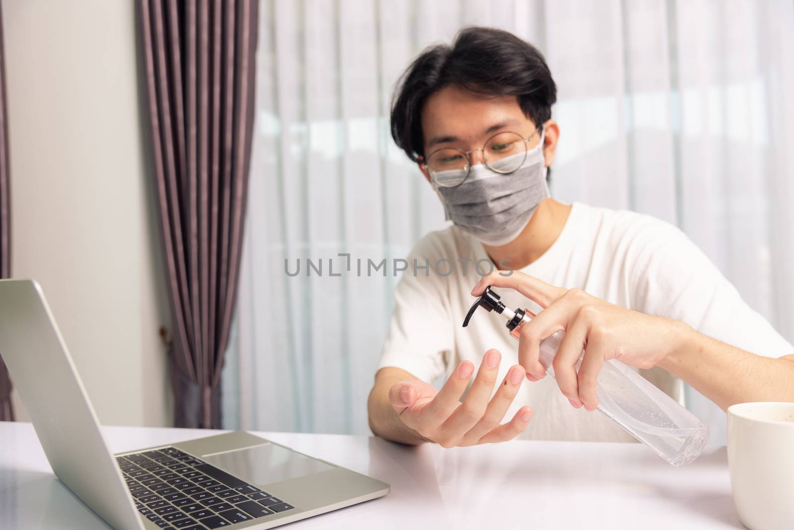 Asian business young handsome man glasses working from home office he quarantines disease coronavirus or COVID-19 wearing protective mask and cleaning hands with sanitizer gel on front laptop computer