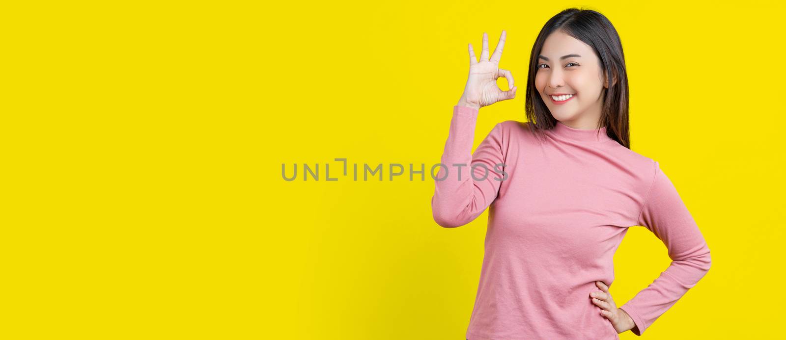 Banner Asian smiling young woman gesturing ok sign for approval or agreement on isolated yellow color background,wearing winter cozy sweater indoors studio,happiness and optimistic decision copy space