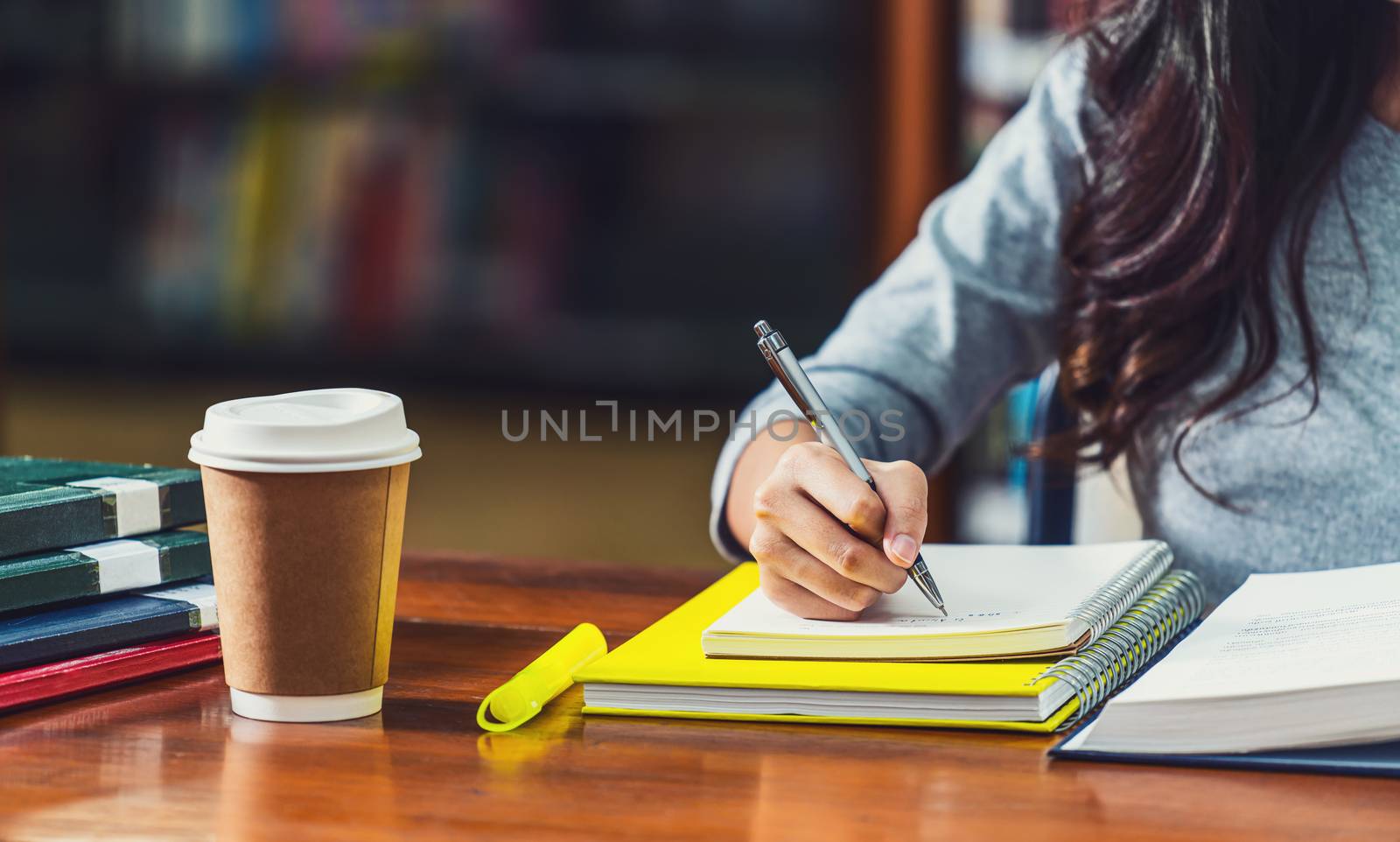 Closeup Asian young Student hand writing homework in library of university or colleague with various book and stationary with coffee cup on wooden table over the book shelf background,Back to school