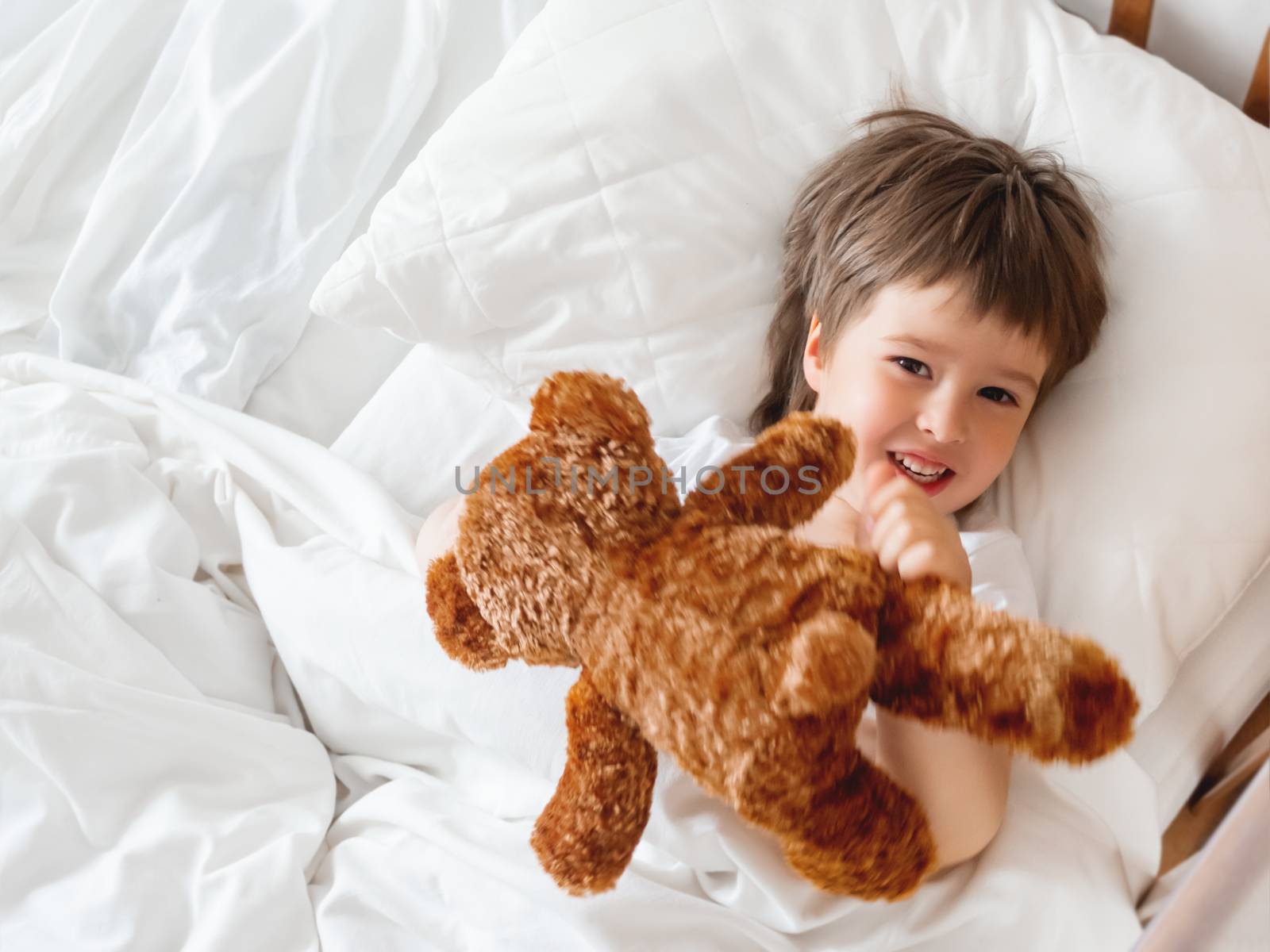 Toddler lies in bed with cute teddy bear. Little boy under white blanket with fluffy toy. Plush guard watches out child's sleep. Morning bedtime at cozy home.