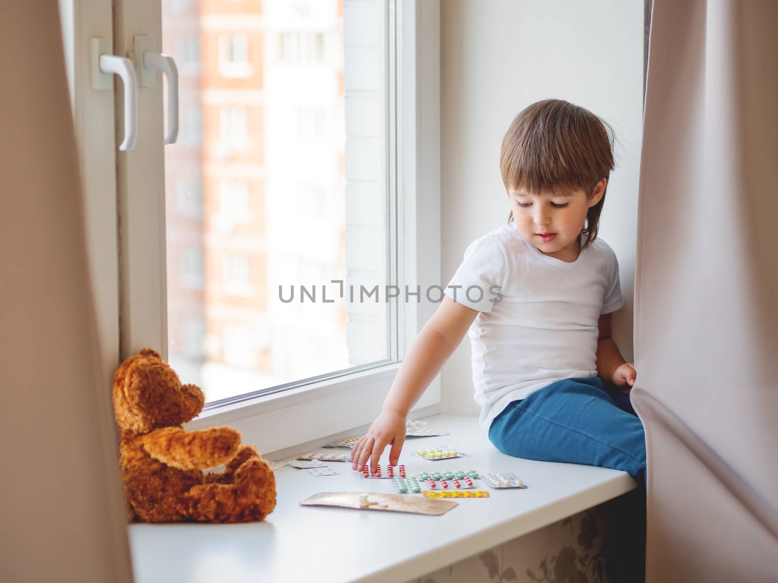 Toddler sits on windowsill and plays with scattering pills without parent's control. Dangerous situation with little boy. Medicines are freely available to child.