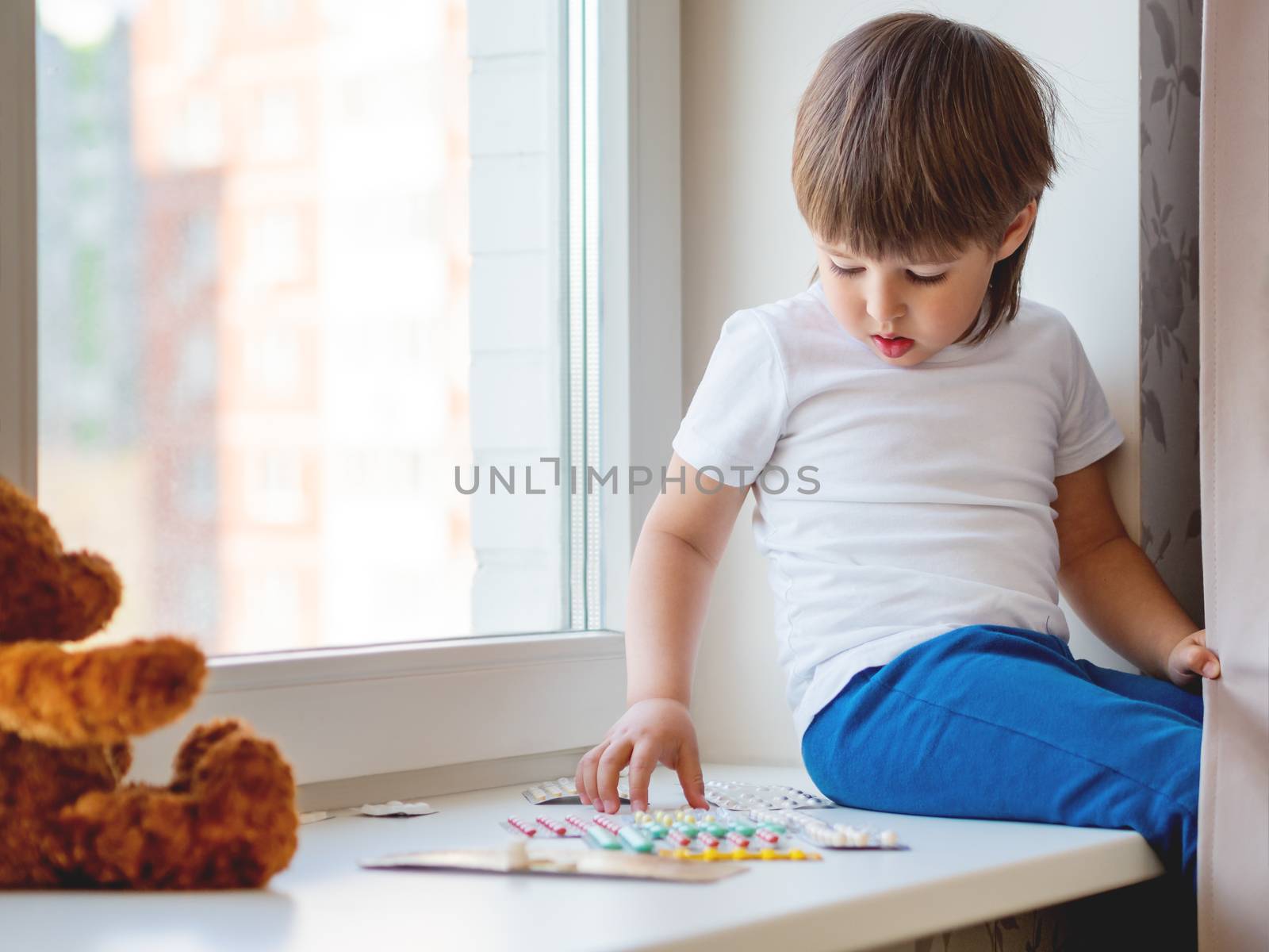 Toddler sits on windowsill and plays with scattering pills without parent's control. Dangerous situation with little boy. Medicines are freely available to child.