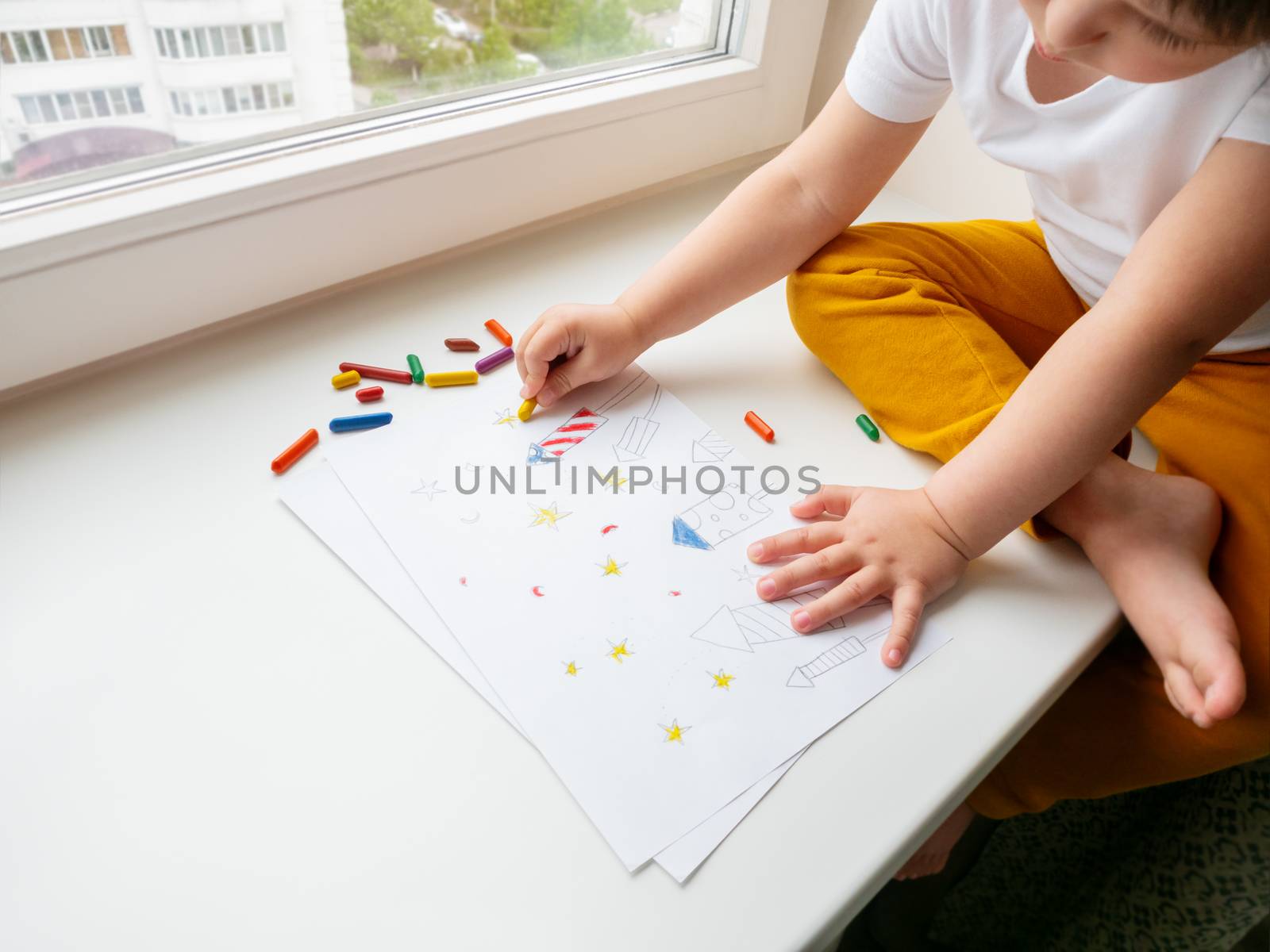 Toddler sits on windowsill and paints colorful fireworks. Child's picture to 4th of July celebration. Independence Day of USA symbol.