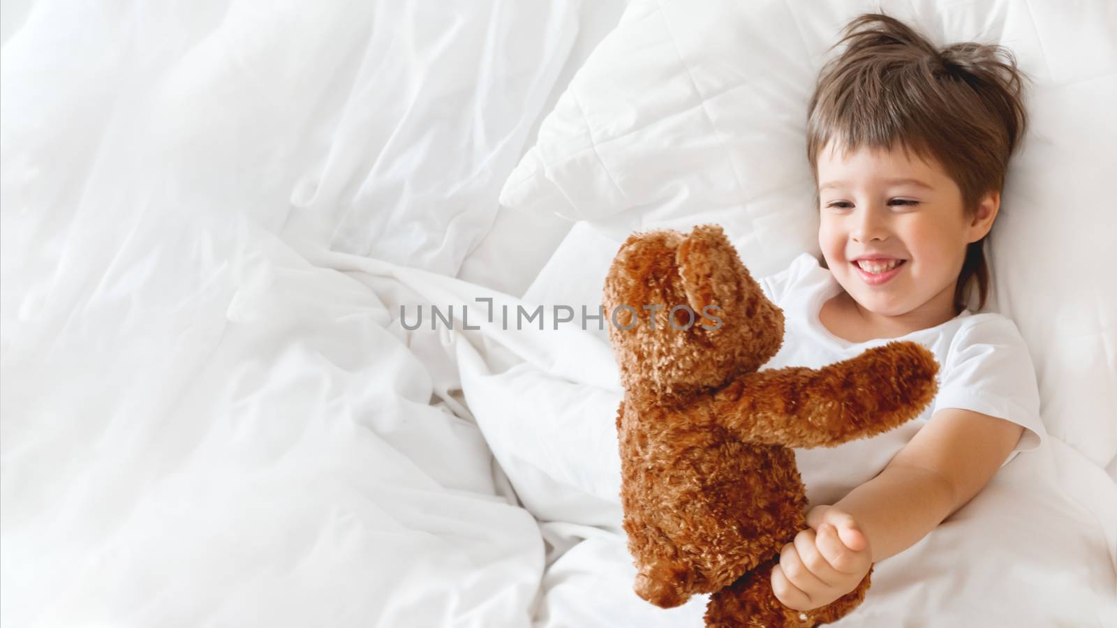 Toddler lies in bed with cute teddy bear. Little boy under white blanket with fluffy toy. Plush guard watches out child's sleep. Morning bedtime at cozy home.