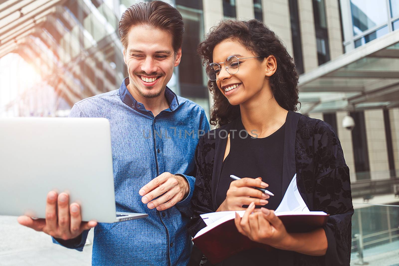 Two smiling colleagues are chatting in the street during lunch break near the building in the background.