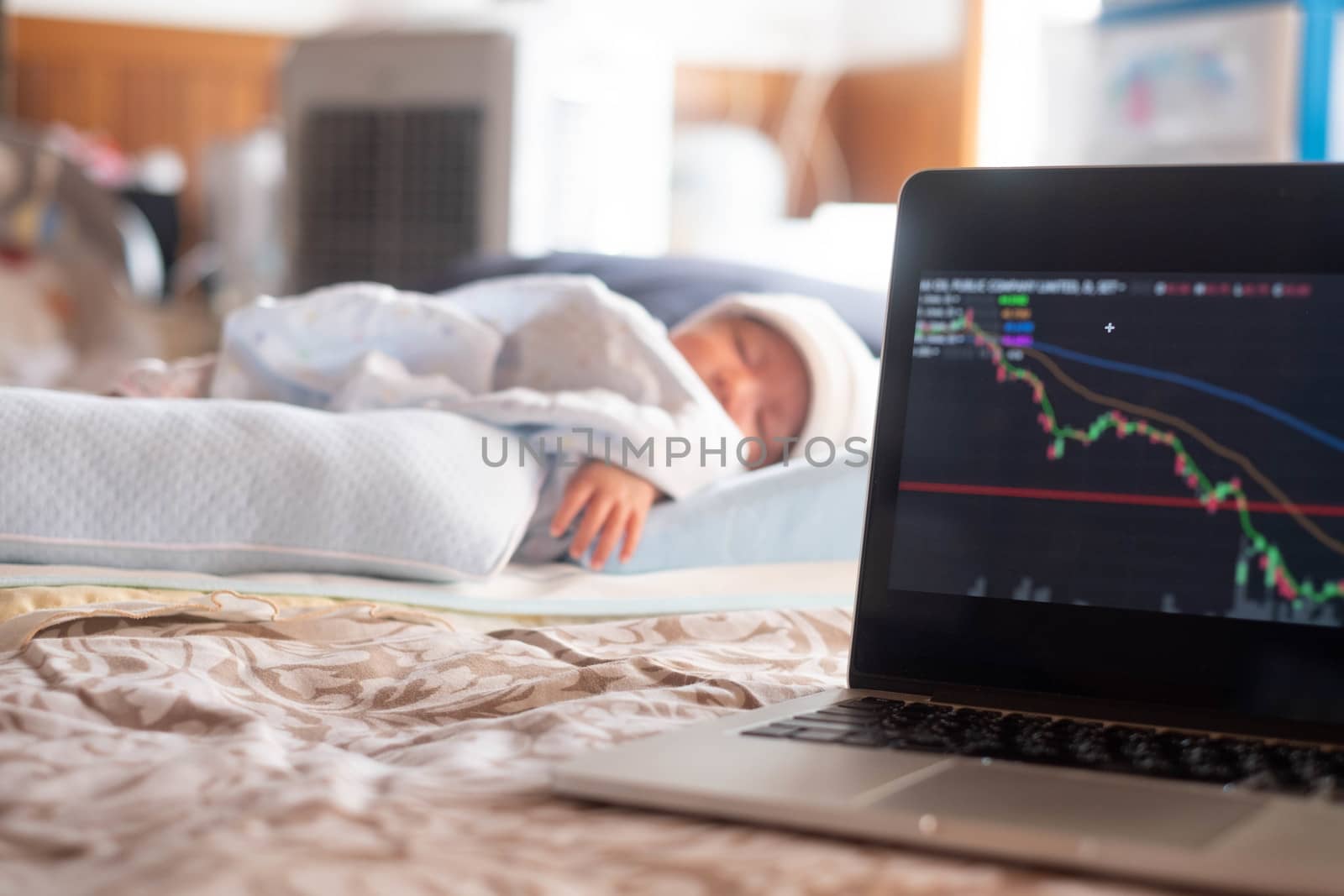 Soft focus on laptop stock display screen .The parents father Stock trader trading stock on his laptop while his newborn infant baby sleep on the bed