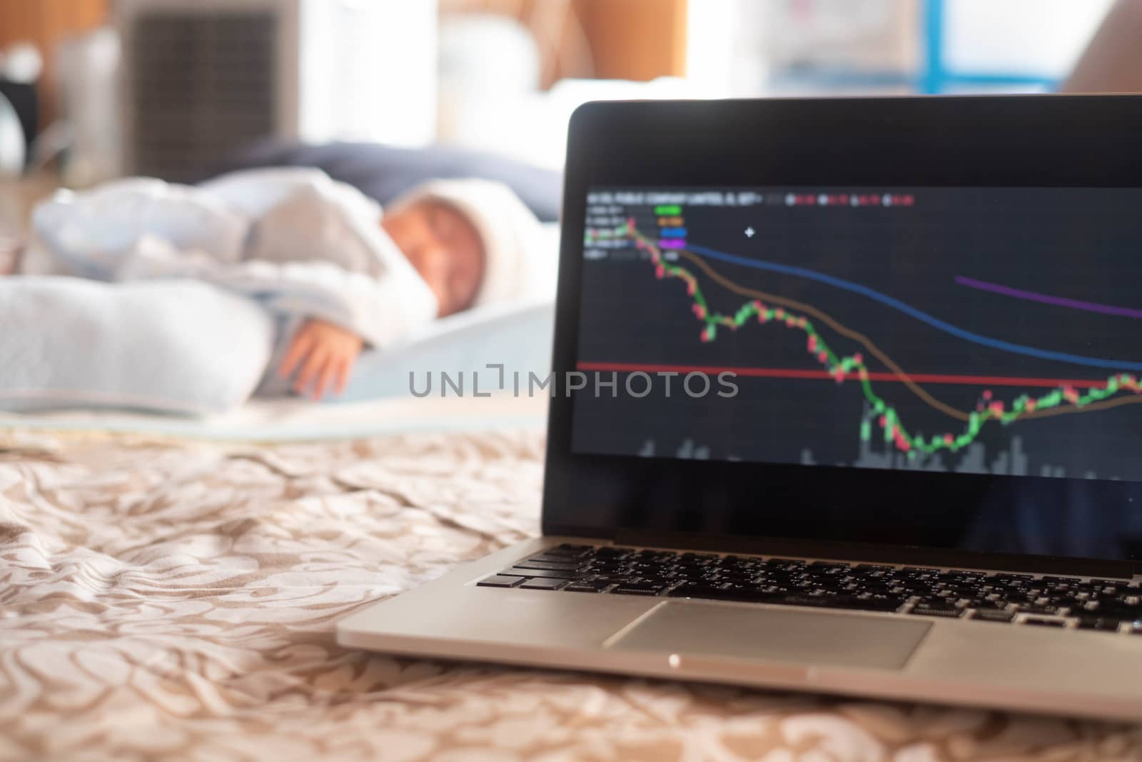 Soft focus on laptop stock display screen .The parents father Stock trader trading stock on his laptop while his newborn infant baby sleep on the bed
