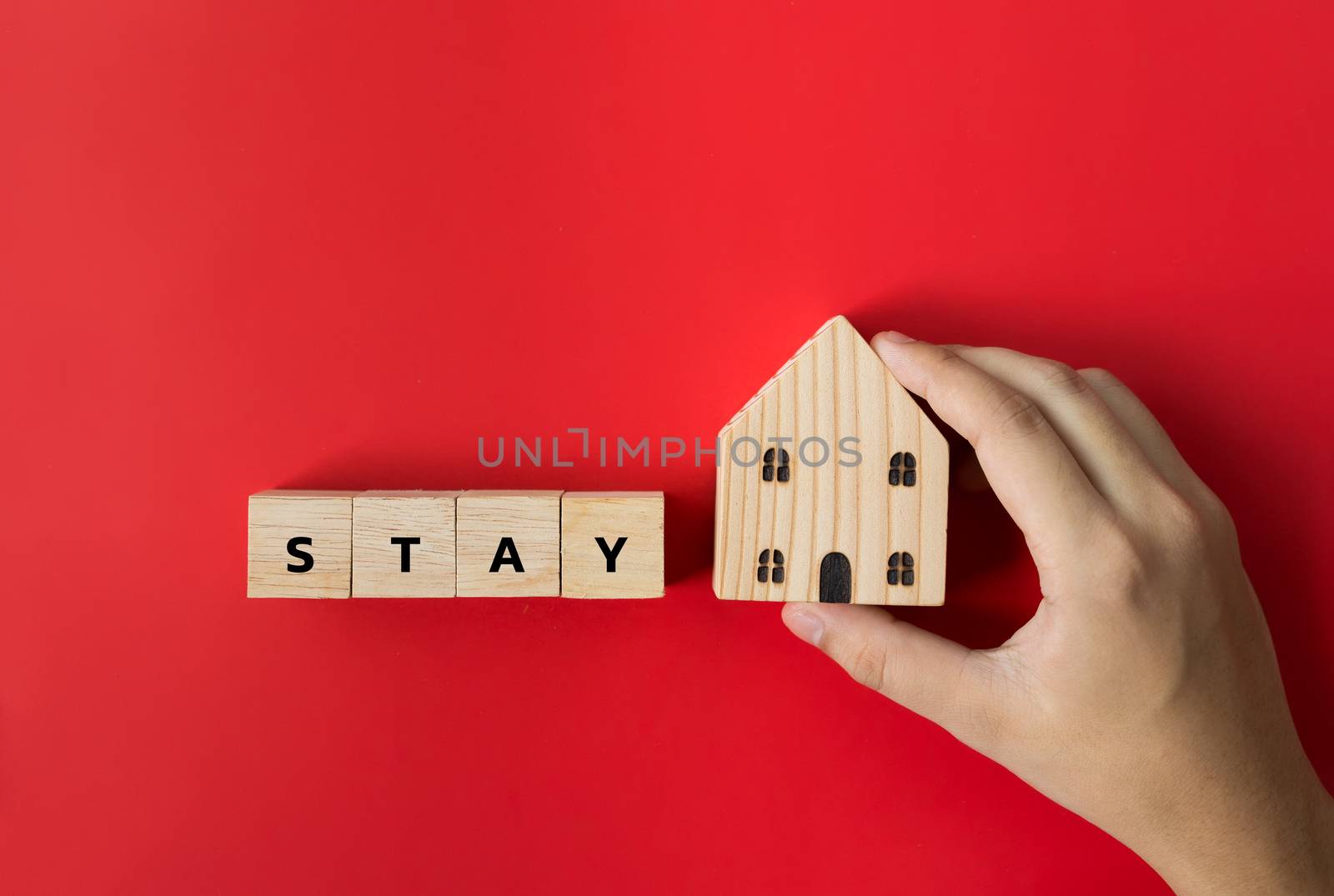 Hand hold on to the house model and text the word "Stay" on the wooden dice. In the concept, stay at home to stop the spread. During the corona virus outbreak or COVID-19 on red background