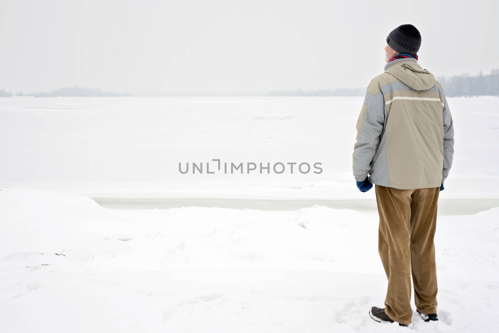 A man with a wool cap, a waterproof jacket and a wool cap is looking at the Dnieper river during a cold and sad gray winter morning under the snow
