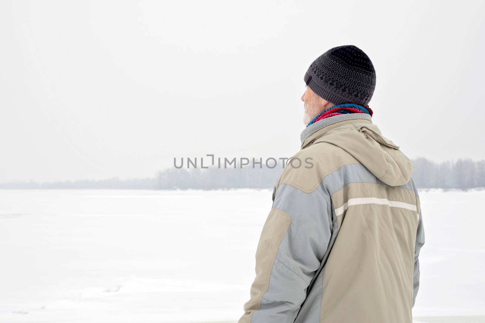 A man with a wool cap, a waterproof jacket and a wool cap is looking at the Dnieper river during a cold and sad gray winter morning under the snow