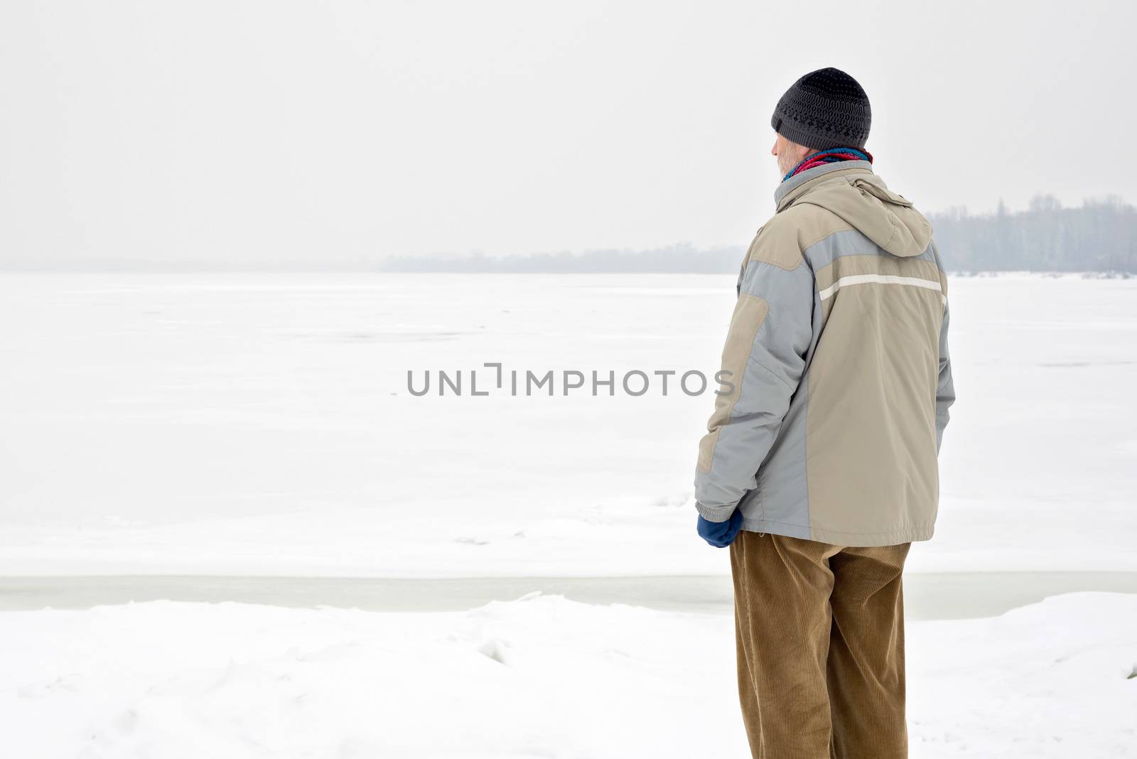 A man with a wool cap, a waterproof jacket and a wool cap is looking at the Dnieper river during a cold and sad gray winter morning under the snow