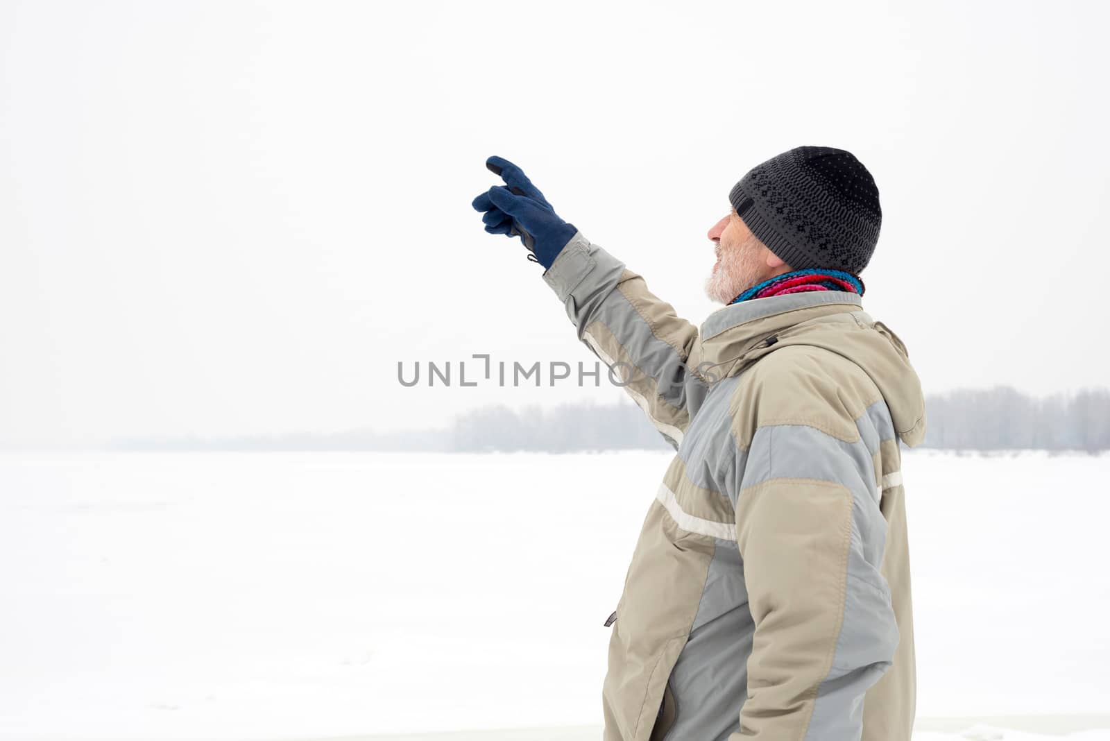 A man with a wool cap, a waterproof jacket and a wool cap is indicating something with the finger close to the Dnieper river during a cold and sad gray winter morning under the snow