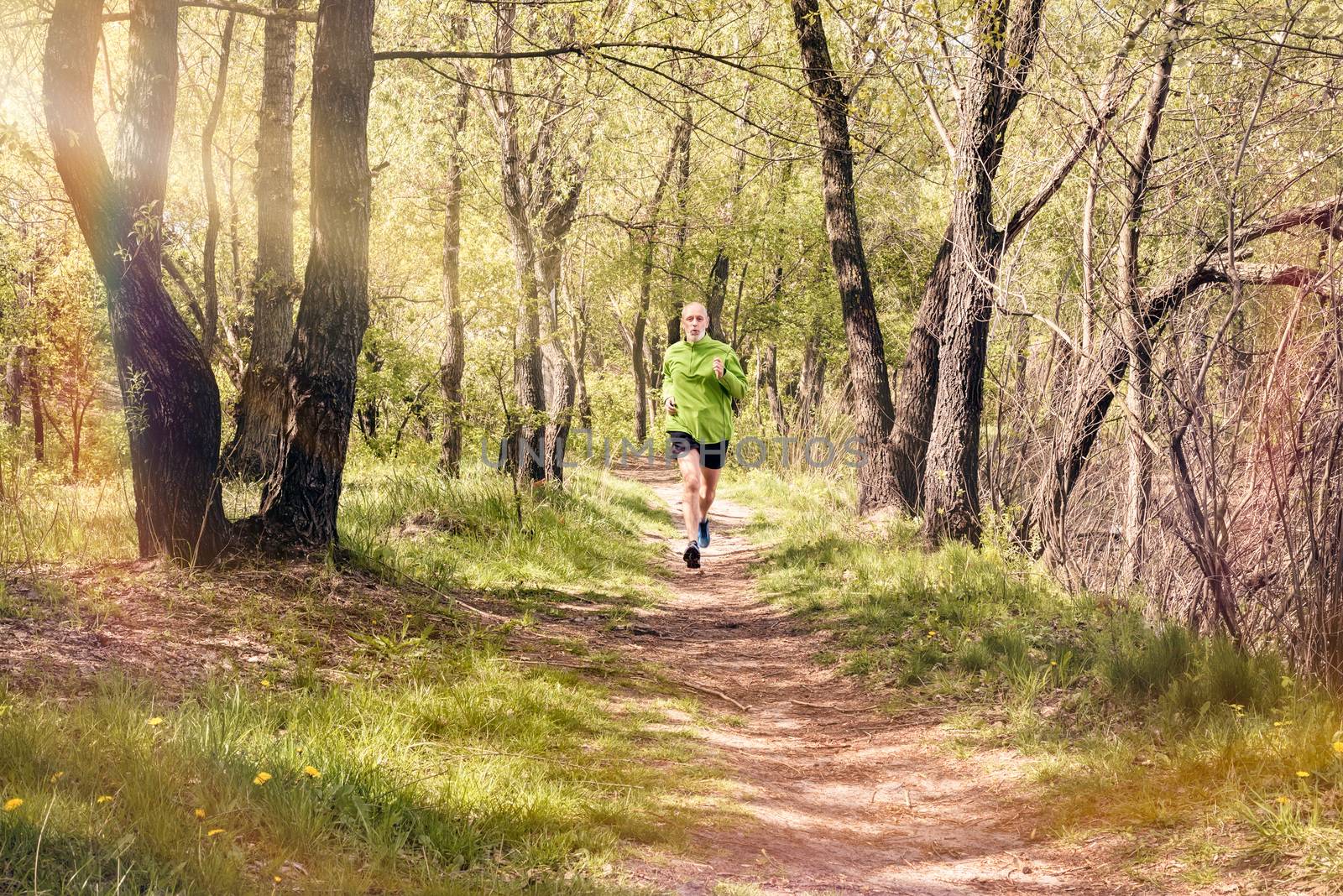 A senior man dressed in black and green is running in the forest, during a warm spring day