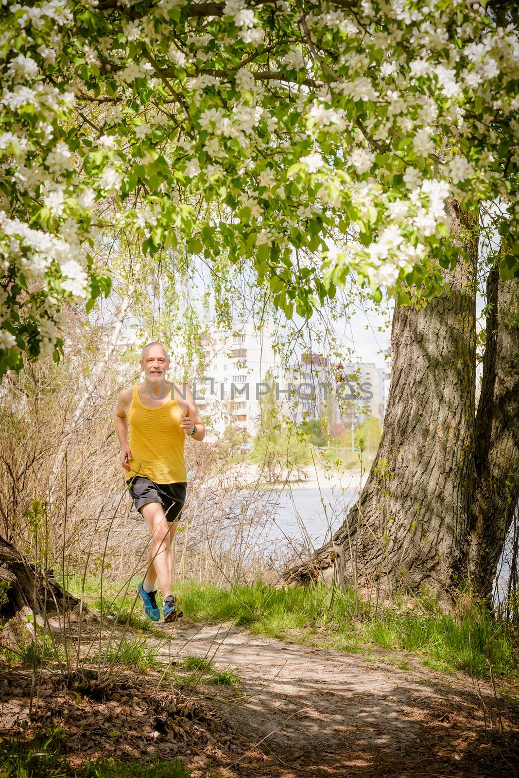 A senior man dressed in black and yellow is running in the forest, close to the lake, during a warm spring day