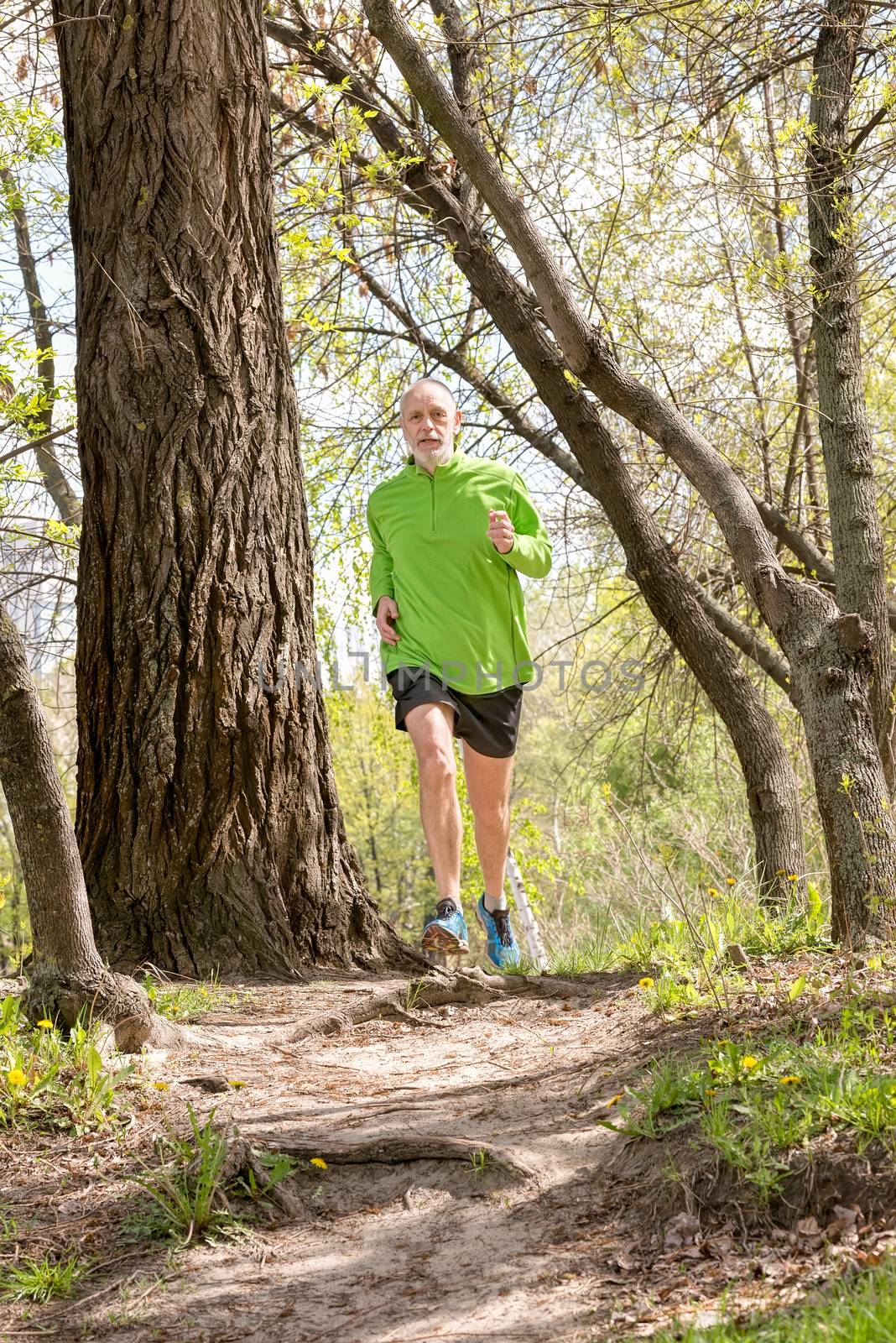 A senior man dressed in black and green is running in the forest, during a warm spring day