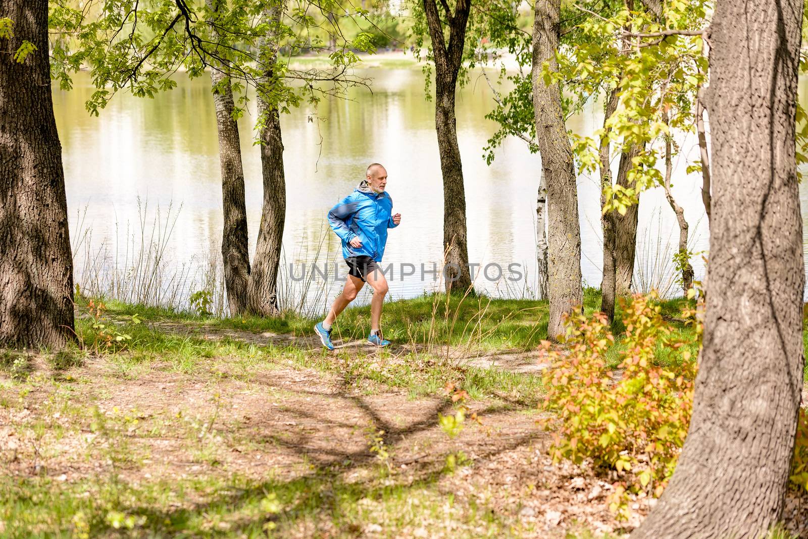 A senior man dressed in black and blue is running close to the lake during a warm spring day