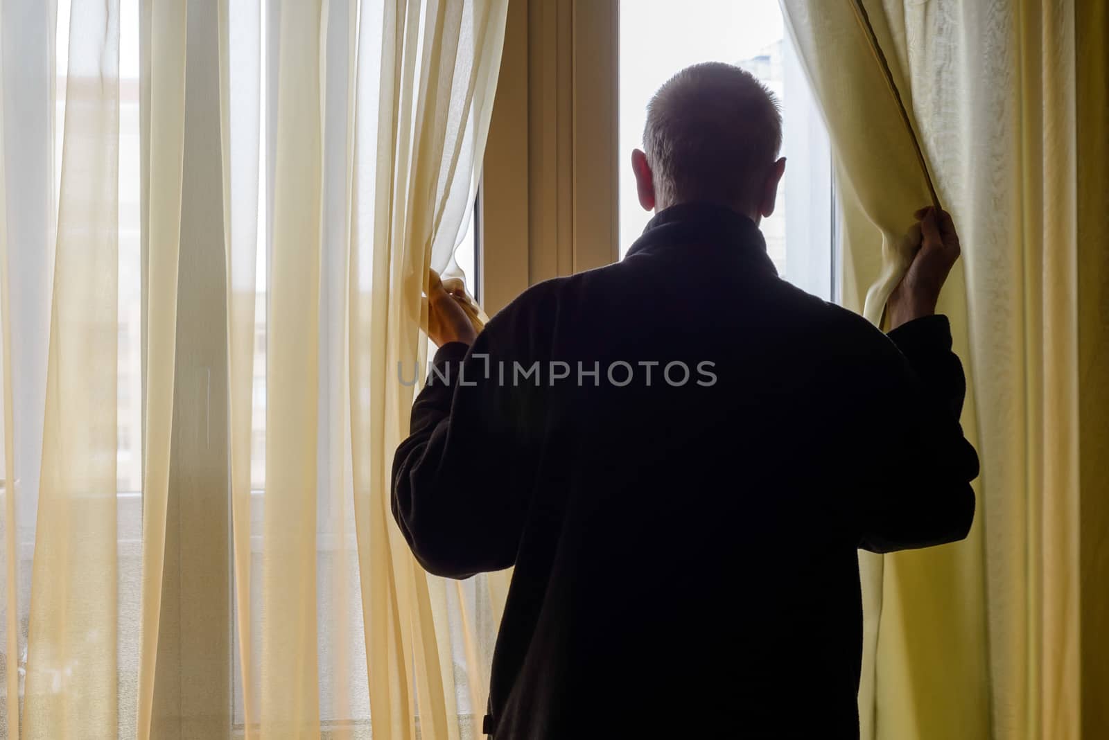 A man in black moves the curtain aside to look out trough the window