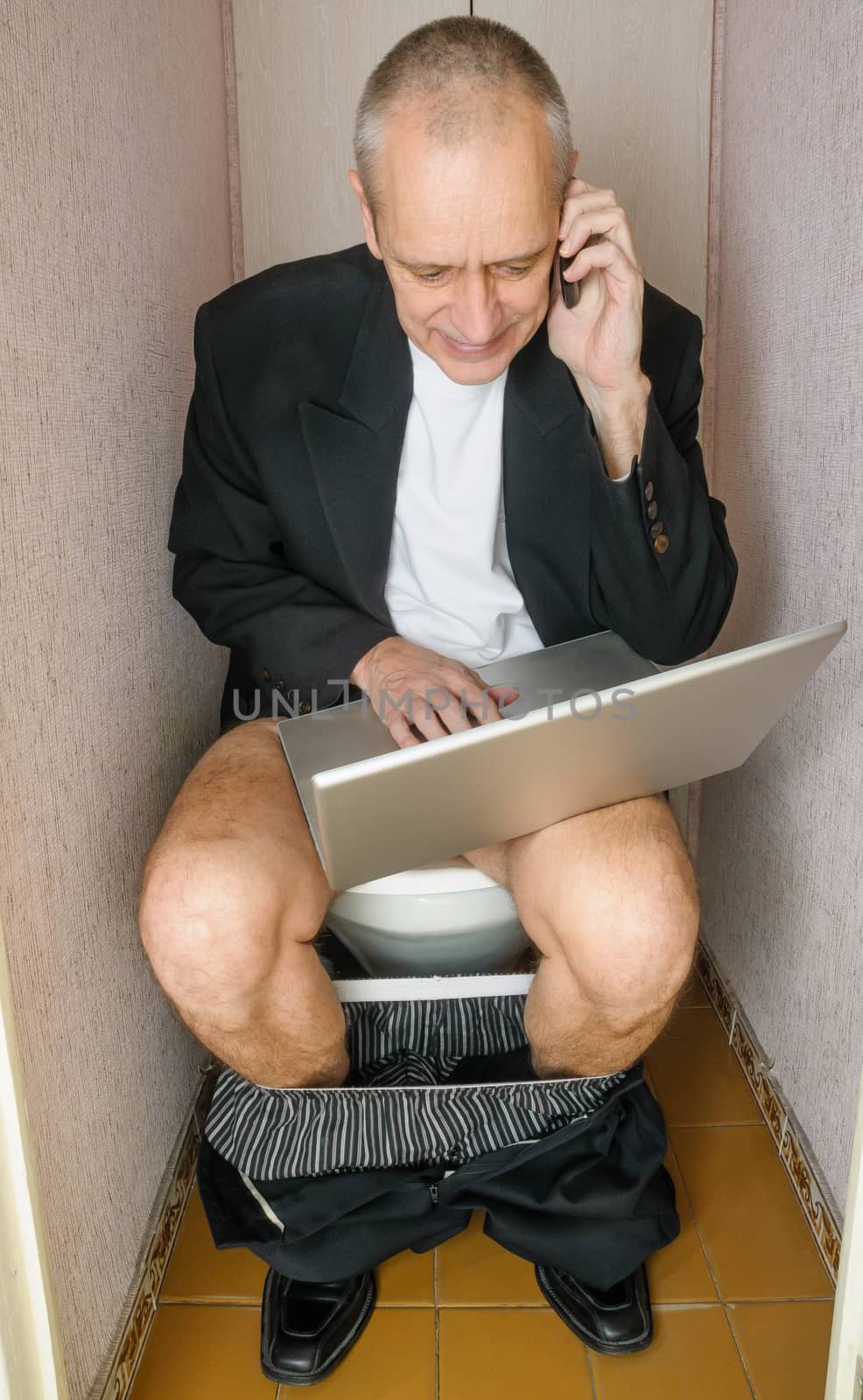 A workaholic adult businessman is using the computer laptop and the mobile phone, also called smartphone, while he is sitting on the bowl in the toilet