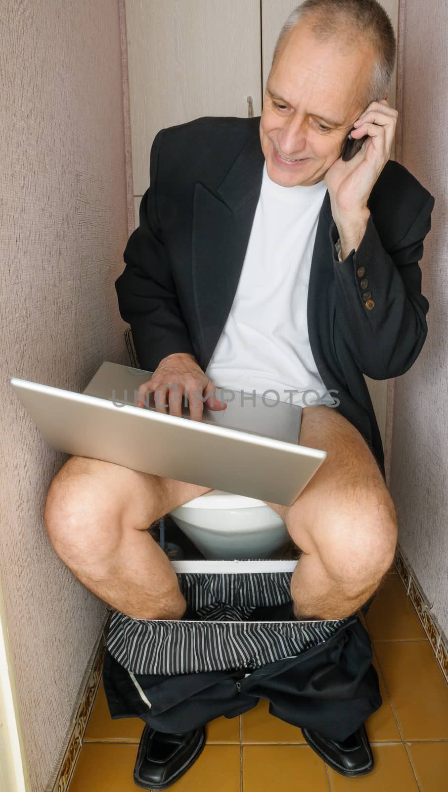 A workaholic adult businessman is using the computer laptop and the mobile phone, also called smartphone, while he is sitting on the bowl in the toilet