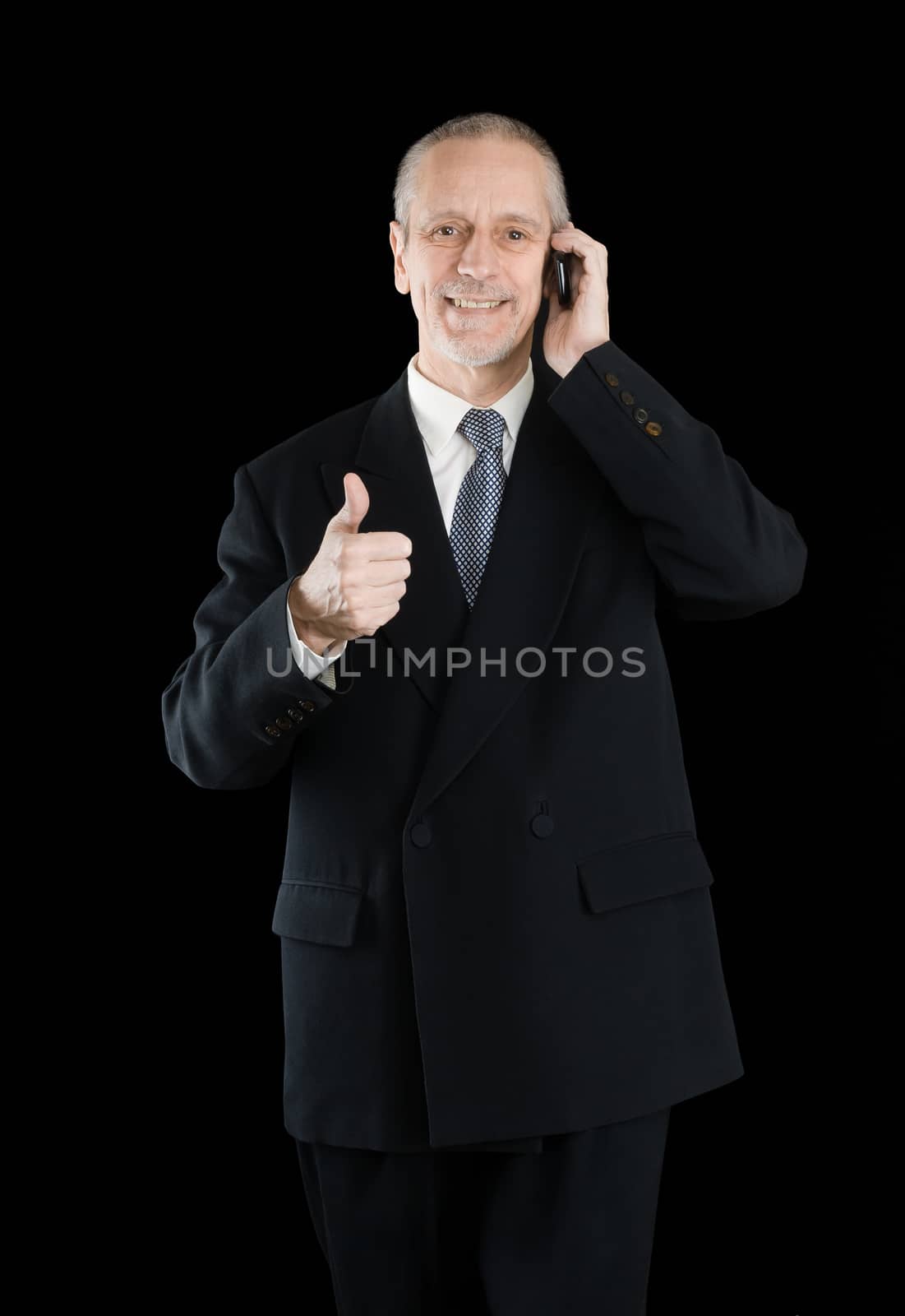 An amiable businessman wearing a black suit  smiling and speaking on mobile phone, with thumb up, on black background