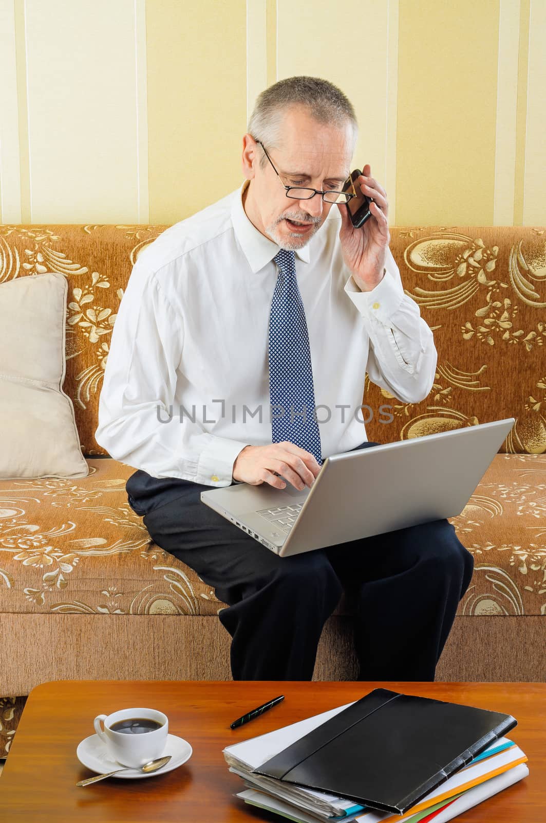An handsome senior businessman working with computer at home, calling to a partner with his mobile phone and ready to drink a hot cup of black coffee
