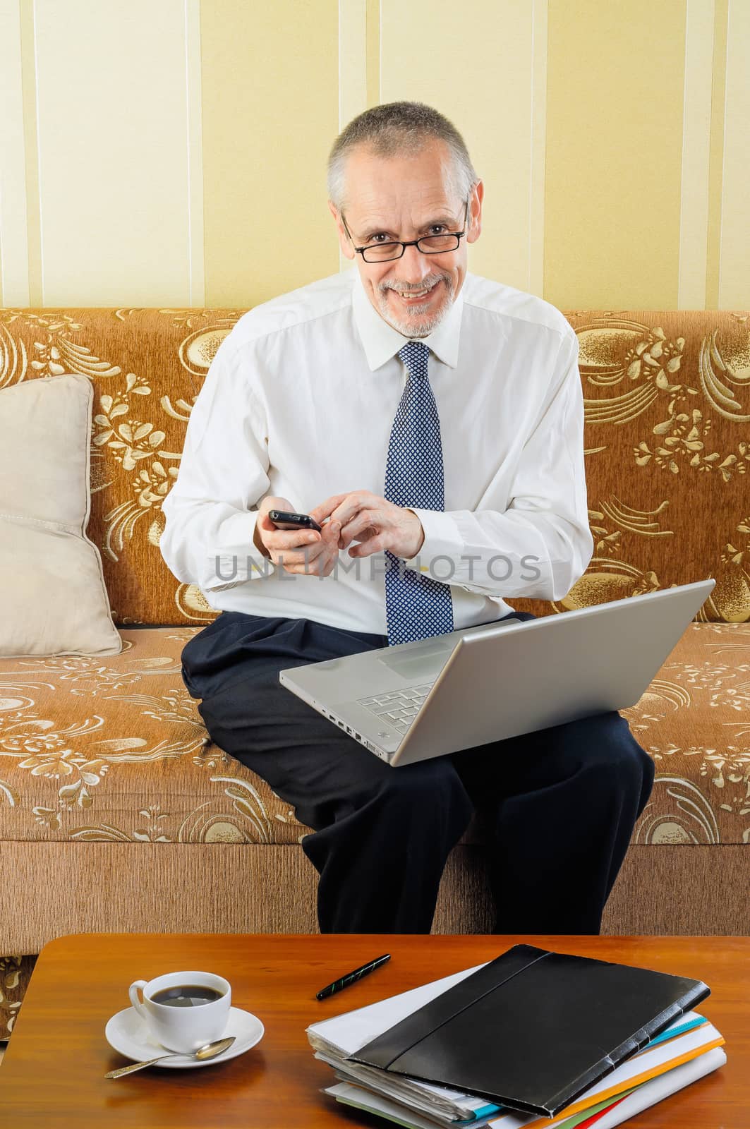 An handsome senior businessman working with computer at home, typing a message on his mobile phone and ready to drink a hot cup of black coffee