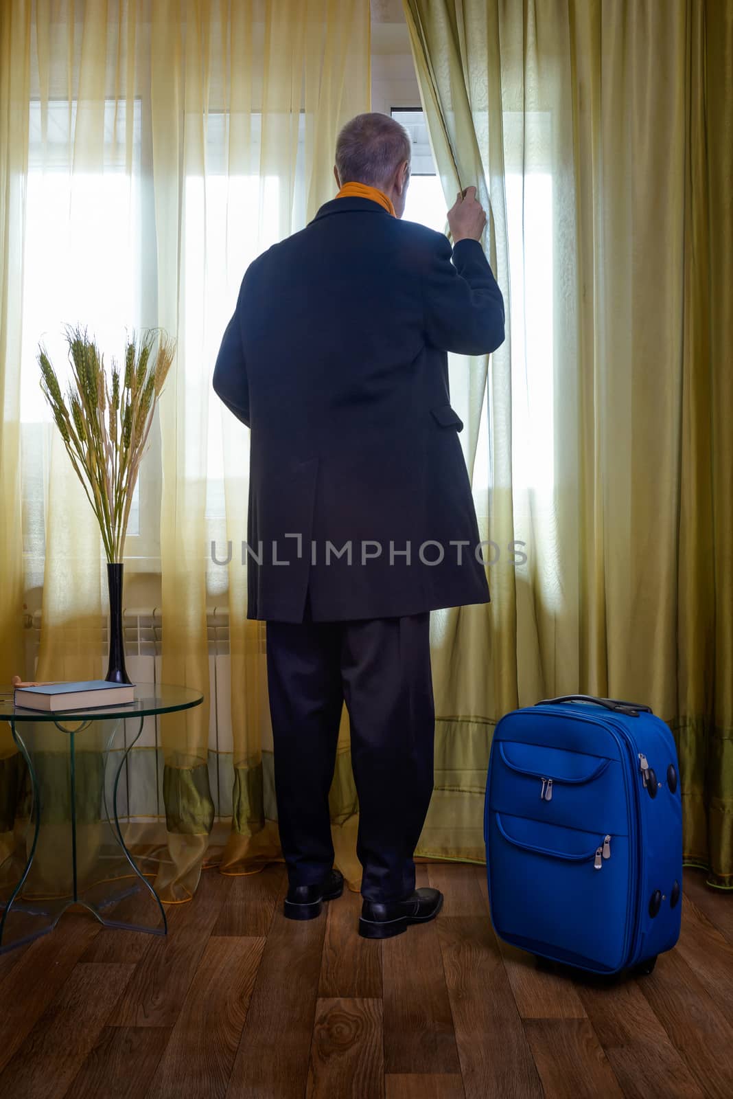 A man in black with a blue suitcase, ready to start for a business travel, moves the curtain aside to look trough the window  if the taxi is coming