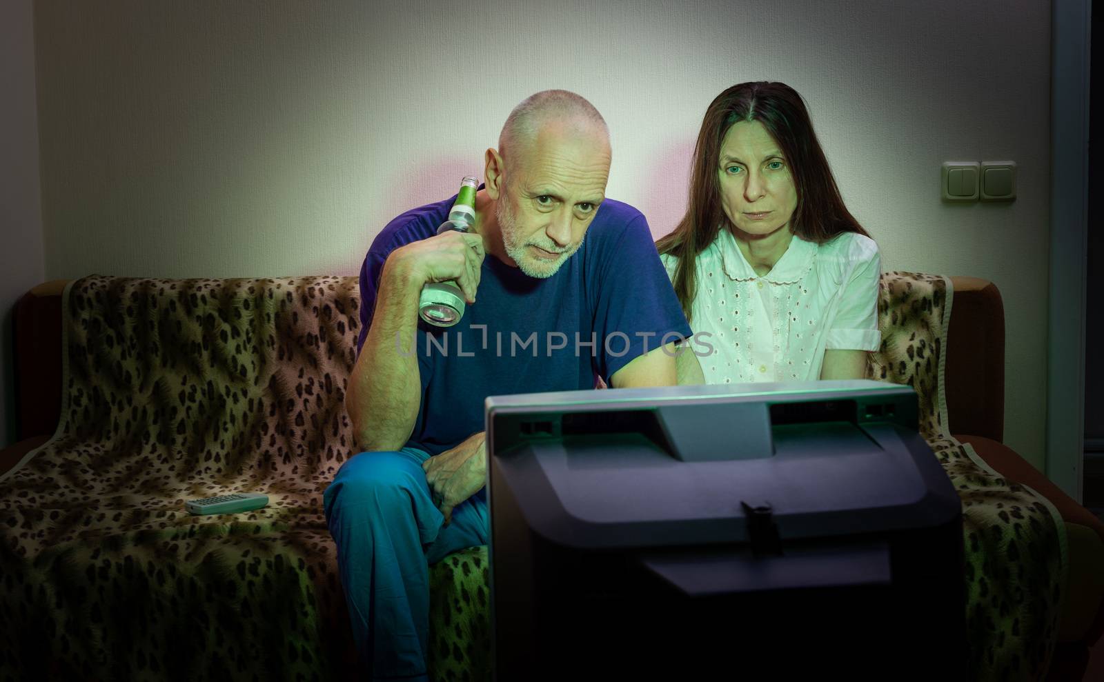 Middle-aged man and woman, watch television, looking sad, sitting on a couch. They watch a sad movie or listen to bad news that makes them a little unhappy and depressed