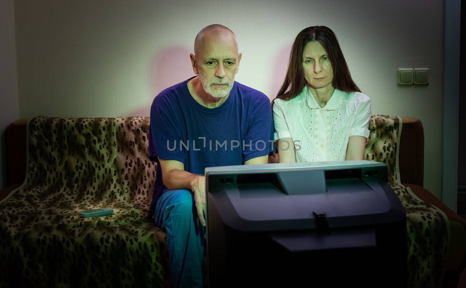 Adult man and woman, watch television, looking sad, sitting on a couch. They watch a sad movie or listen to bad news that makes them a little unhappy and depressed