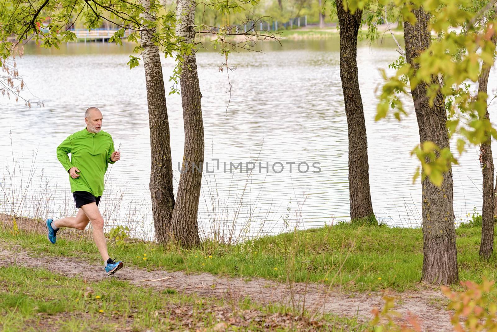 A senior man dressed in black and green is running in the park, close to the lake, during a gray spring day