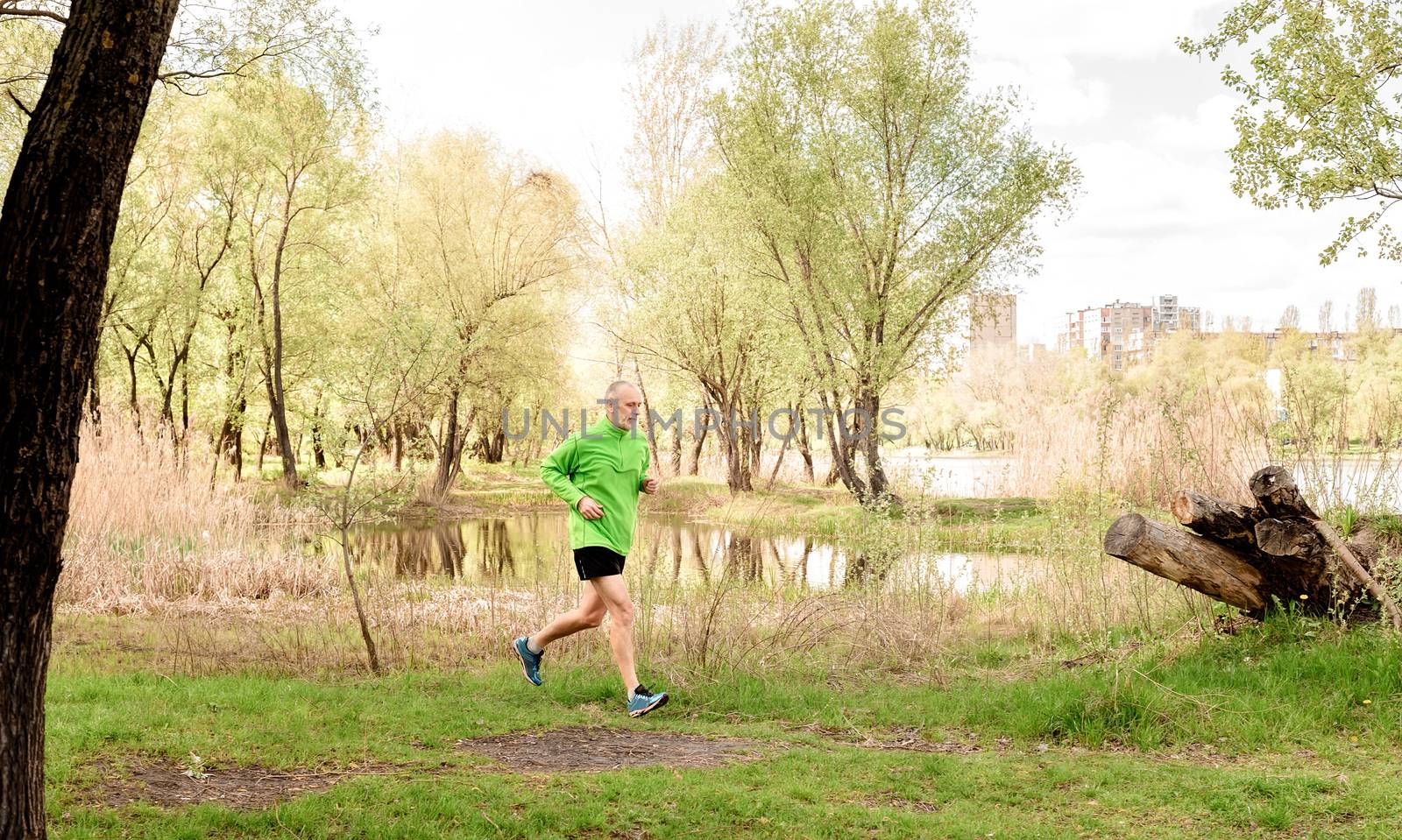 A senior man dressed in black and green is running close to the lake during a fresh spring day