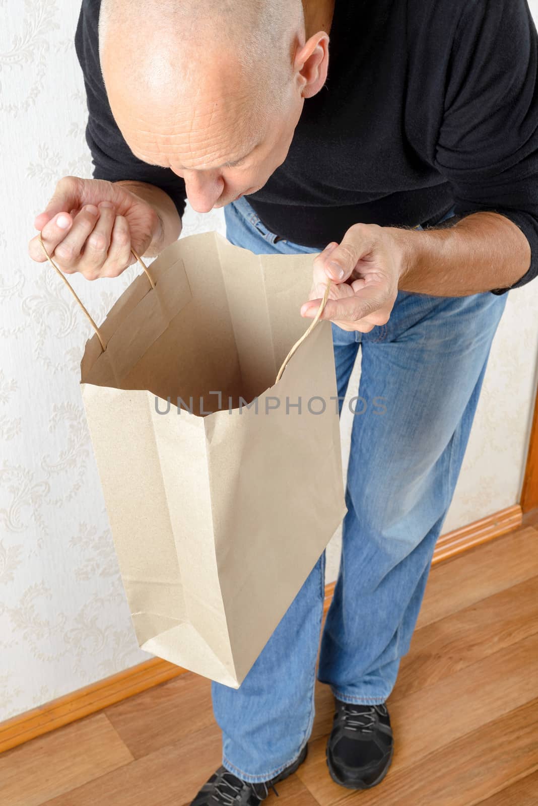 A man in blue jeans and black sweatshirt is looking inside a brown paper bag