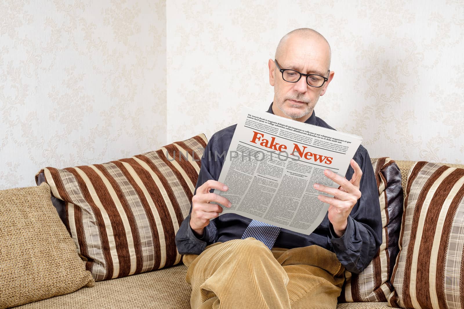 A man wearing glasses is sitting on a couch at home, reading a newspaper reporting fake news. Fake Lorem ipsum text.