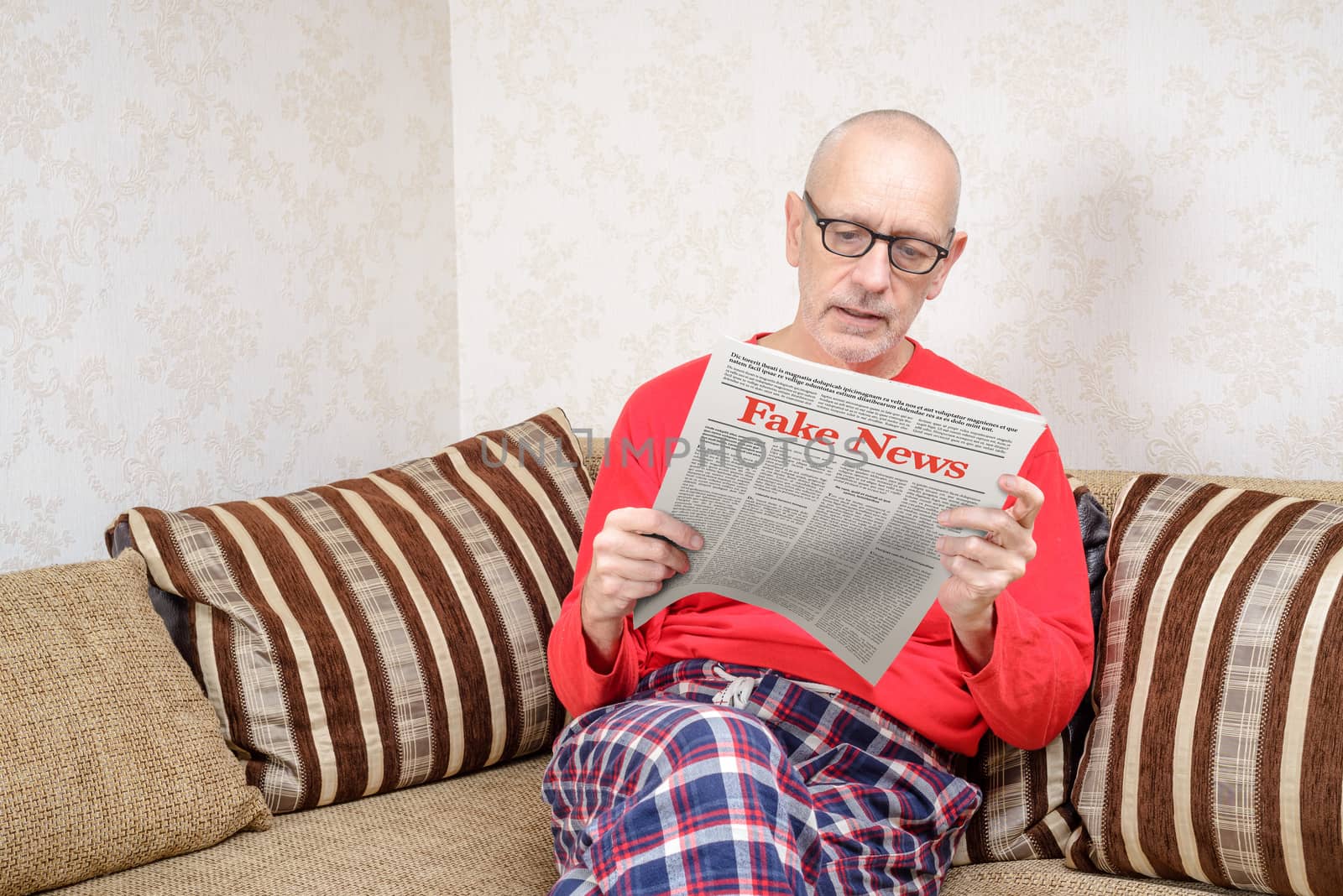 A man wearing glasses and pajamas is sitting on a couch at home, reading a newspaper reporting fake news. Fake Lorem ipsum text.