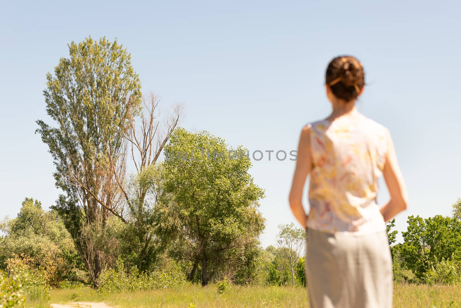 A woman with a chignon, standing up close to the country road in Kiev, Ukraine, observes the trees in the distance. The silhouette of the lady is out of focus, against a focused background.