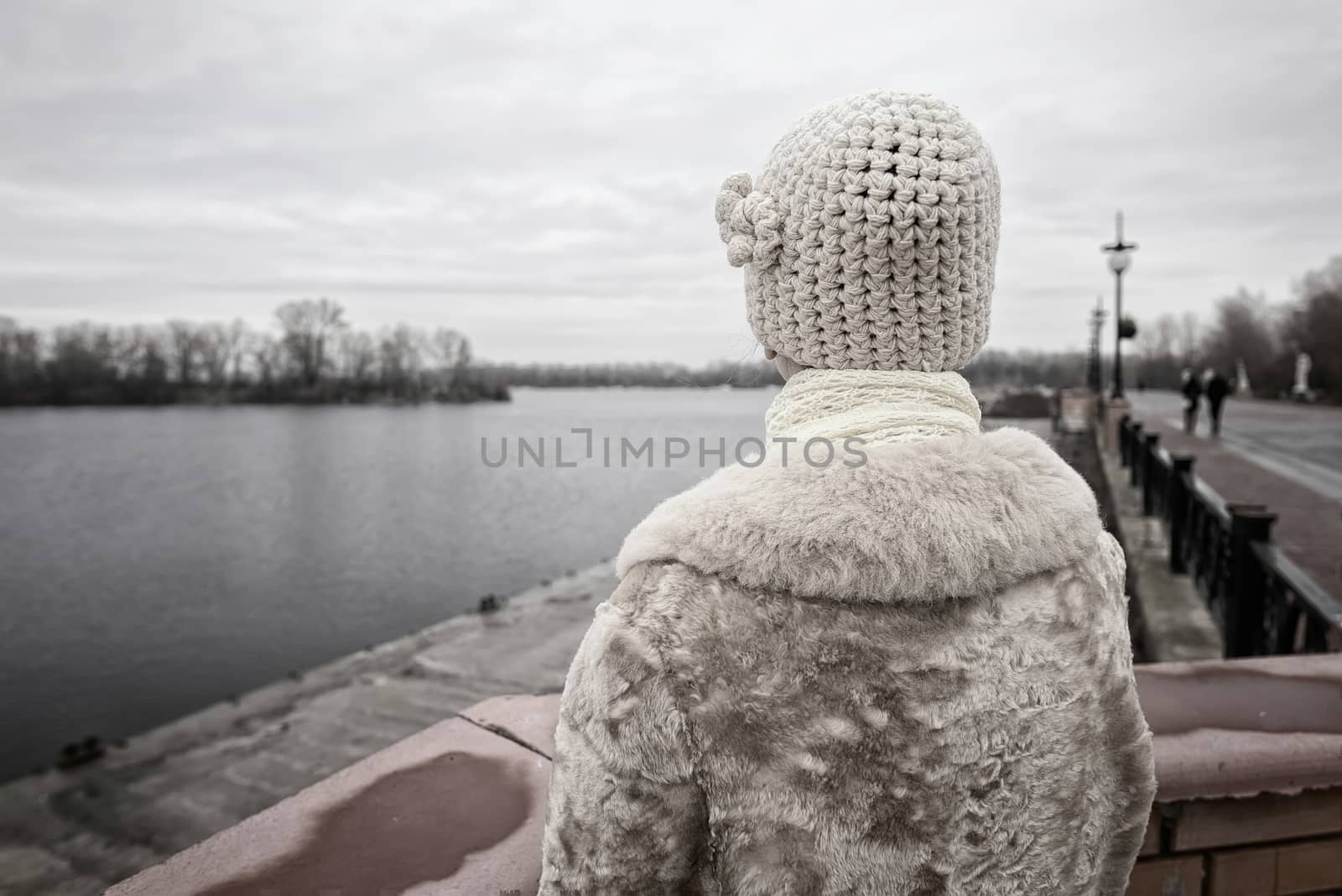 A woman with a wool cap and a fur coat is looking at the river during a cold and sad gray winter morning