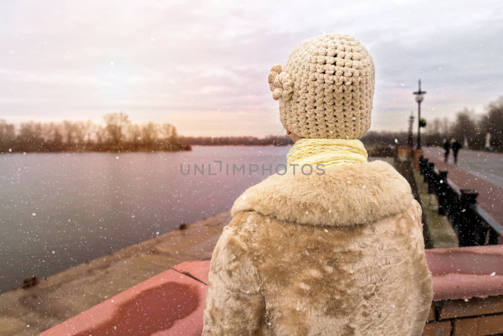 A woman with a wool cap and a fur coat is looking at the river during a cold and sad gray winter morning under the snow