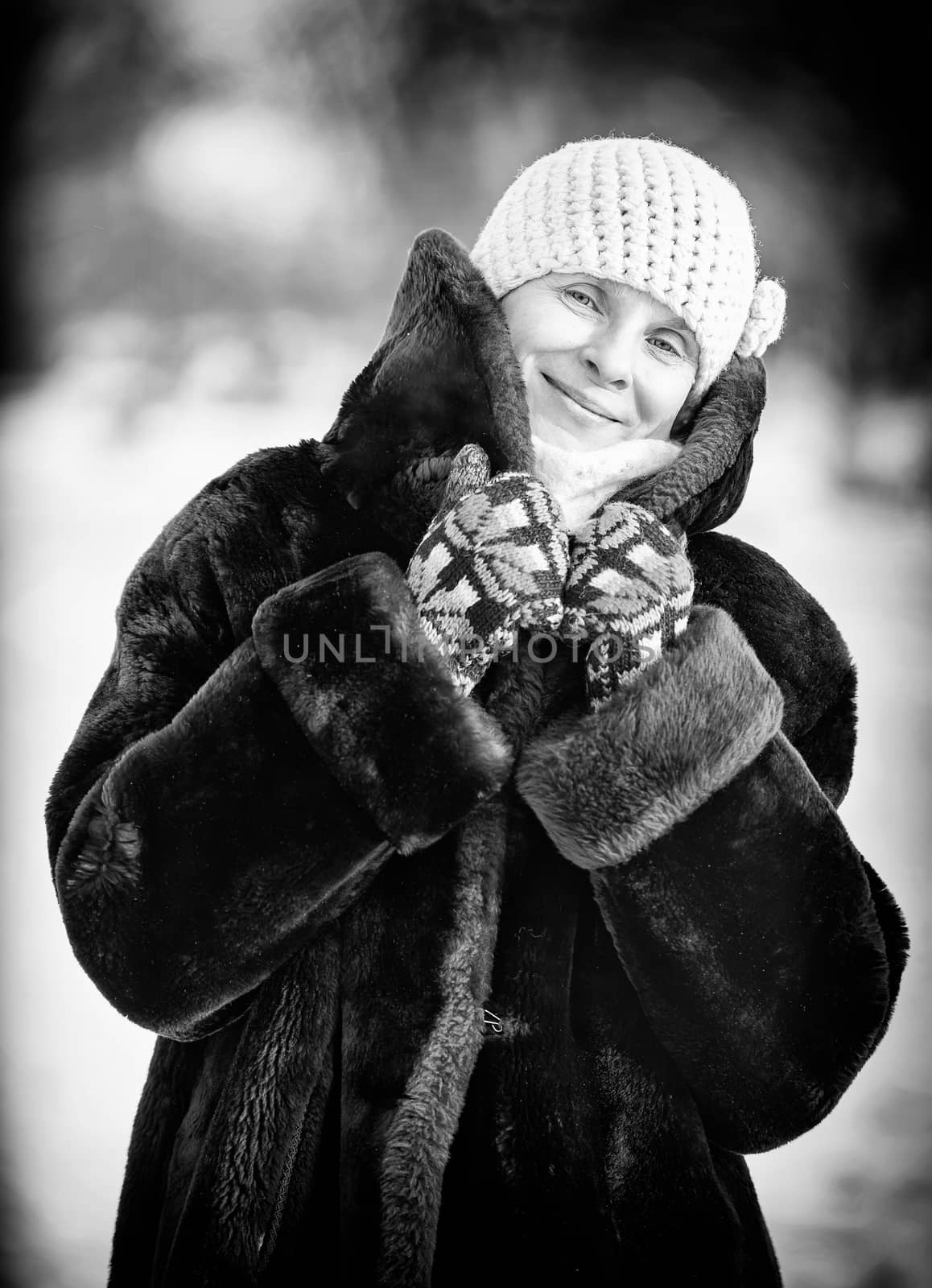 A winter portrait of a smiling senior adult woman wearing a wool cap, a scarf and colored gloves, with a snow background