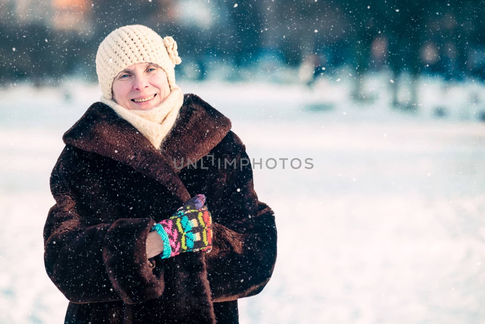 A winter portrait of a smiling senior adult woman wearing a wool cap, a scarf and colored gloves, with a snow background