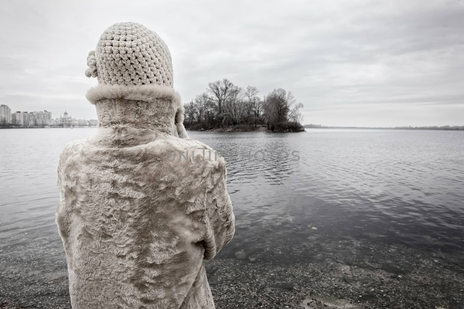 A woman with a wool cap and a fur coat is looking at the river during a cold and sad gray winter morning