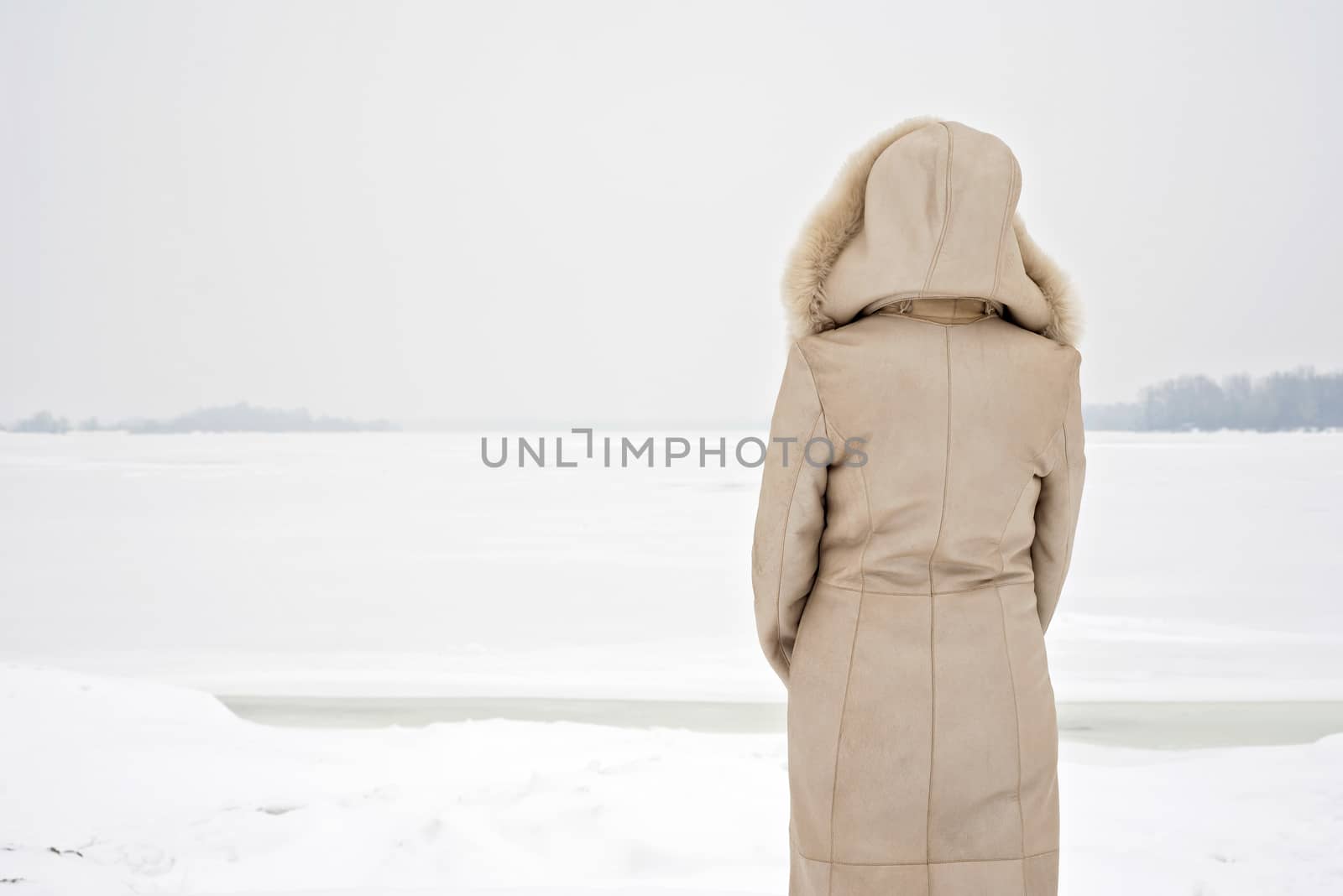 A woman with a leather coat with a fur hood is looking at the frozen Dnieper river during a cold and sad gray winter morning under the snow