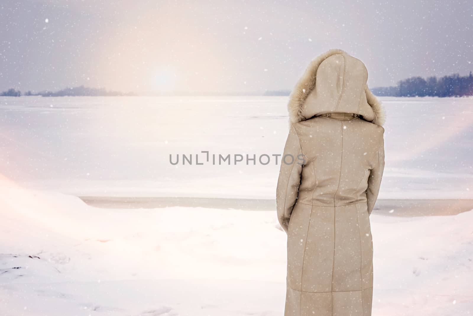 A woman with a leather coat with a fur hood is looking at the frozen Dnieper river during a cold and sad gray winter morning under the snow