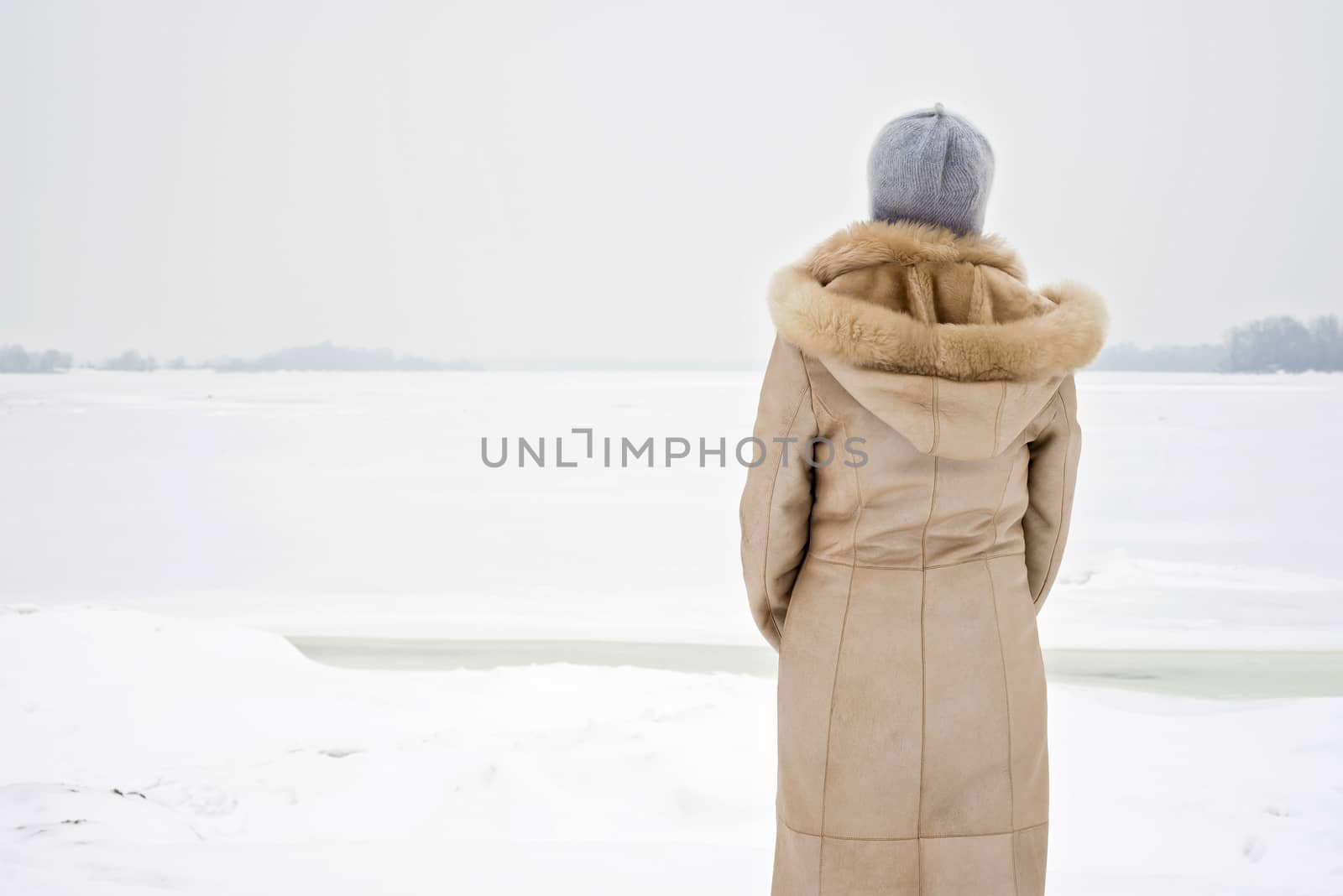 A woman with a wool cap and a leather coat is looking at the Dnieper river during a cold and sad gray winter morning under the snow